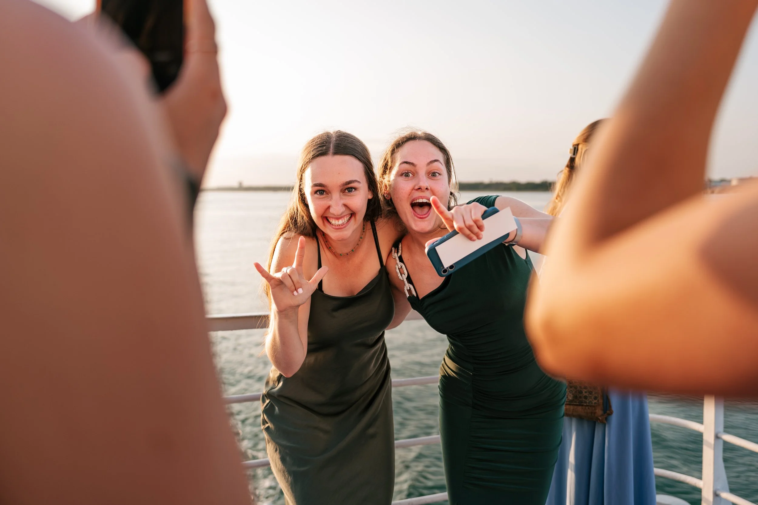 Two women in black dresses taking a selfie on a boat, smiling and making peace signs, with water and a distant shoreline in the background.