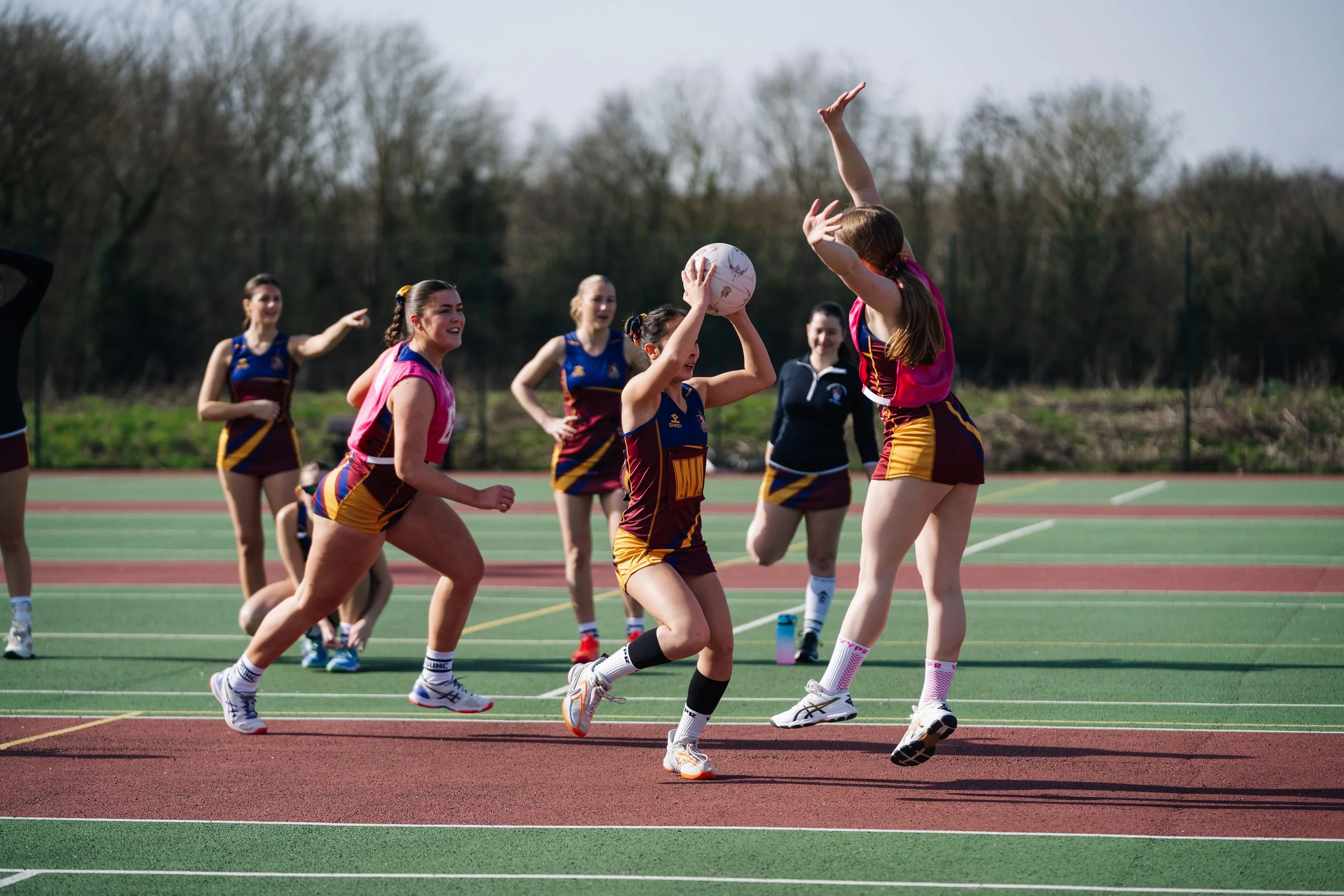 Girls playing netball on an outdoor court, with one girl jumping to catch the ball while others watch and support.