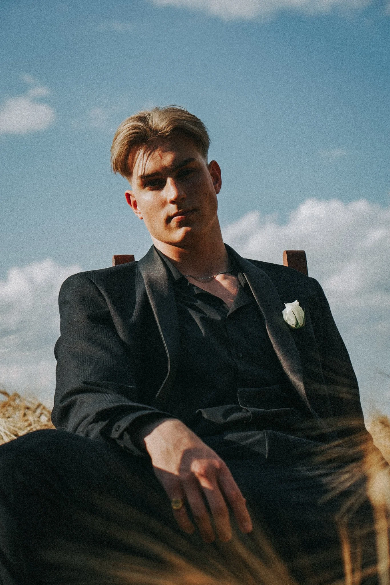 Young man in black suit with a white boutonniere sitting outdoors under a partly cloudy sky, with a serious expression.