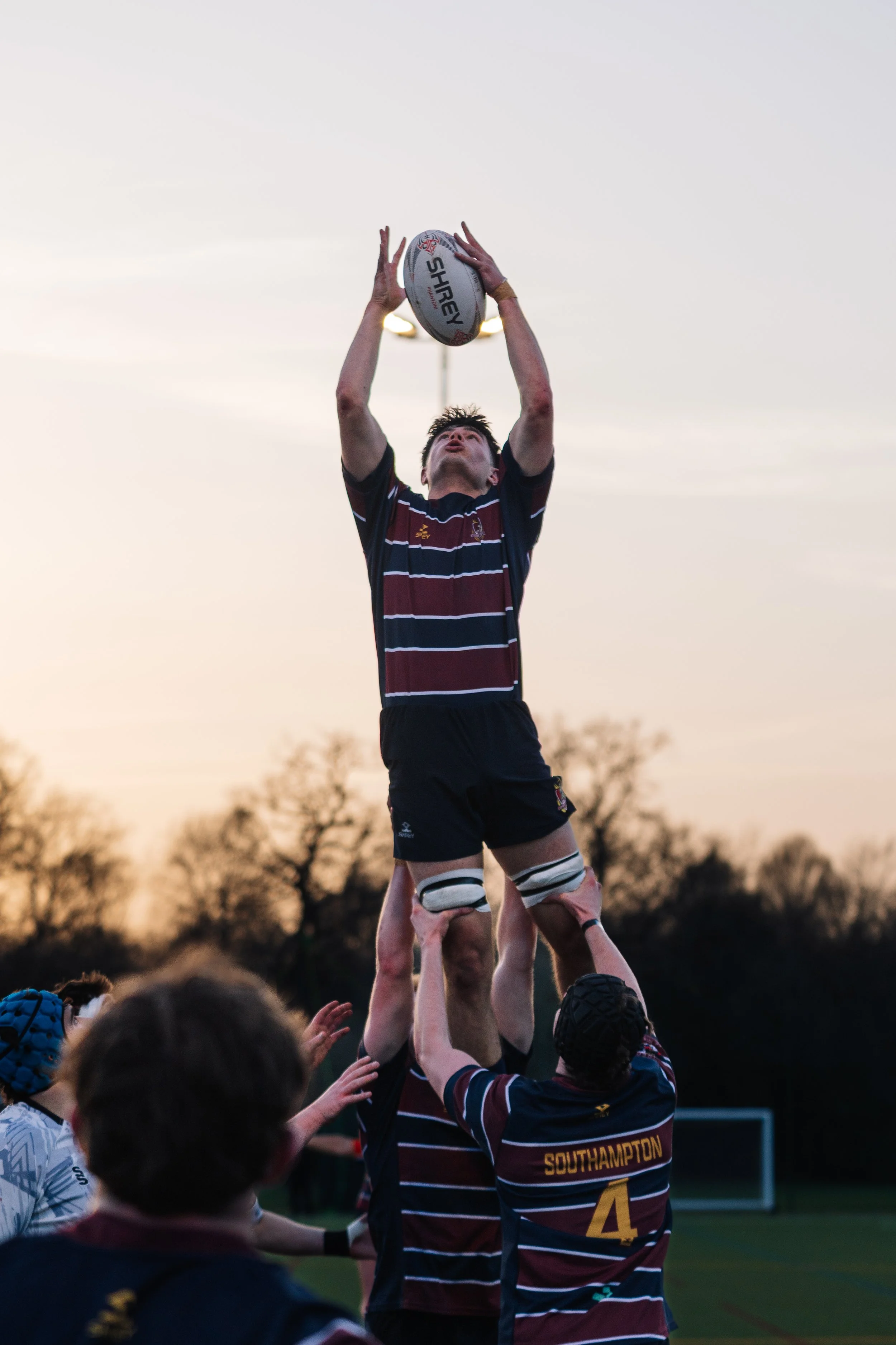 Rugby players perform a line-out catch during a game at sunset, with the sky and trees in the background.