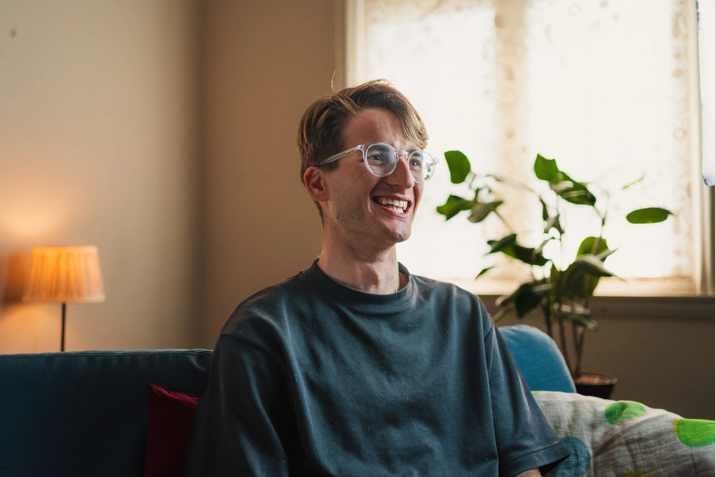 A young man with glasses and a black t-shirt sitting on a blue couch, smiling in a well-lit living room with a potted plant and a lamp in the background.