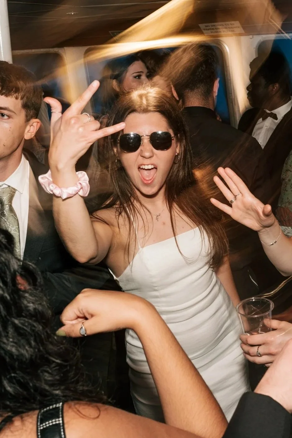 A woman wearing sunglasses and a white dress at a party, making a rock on hand gesture and smiling.