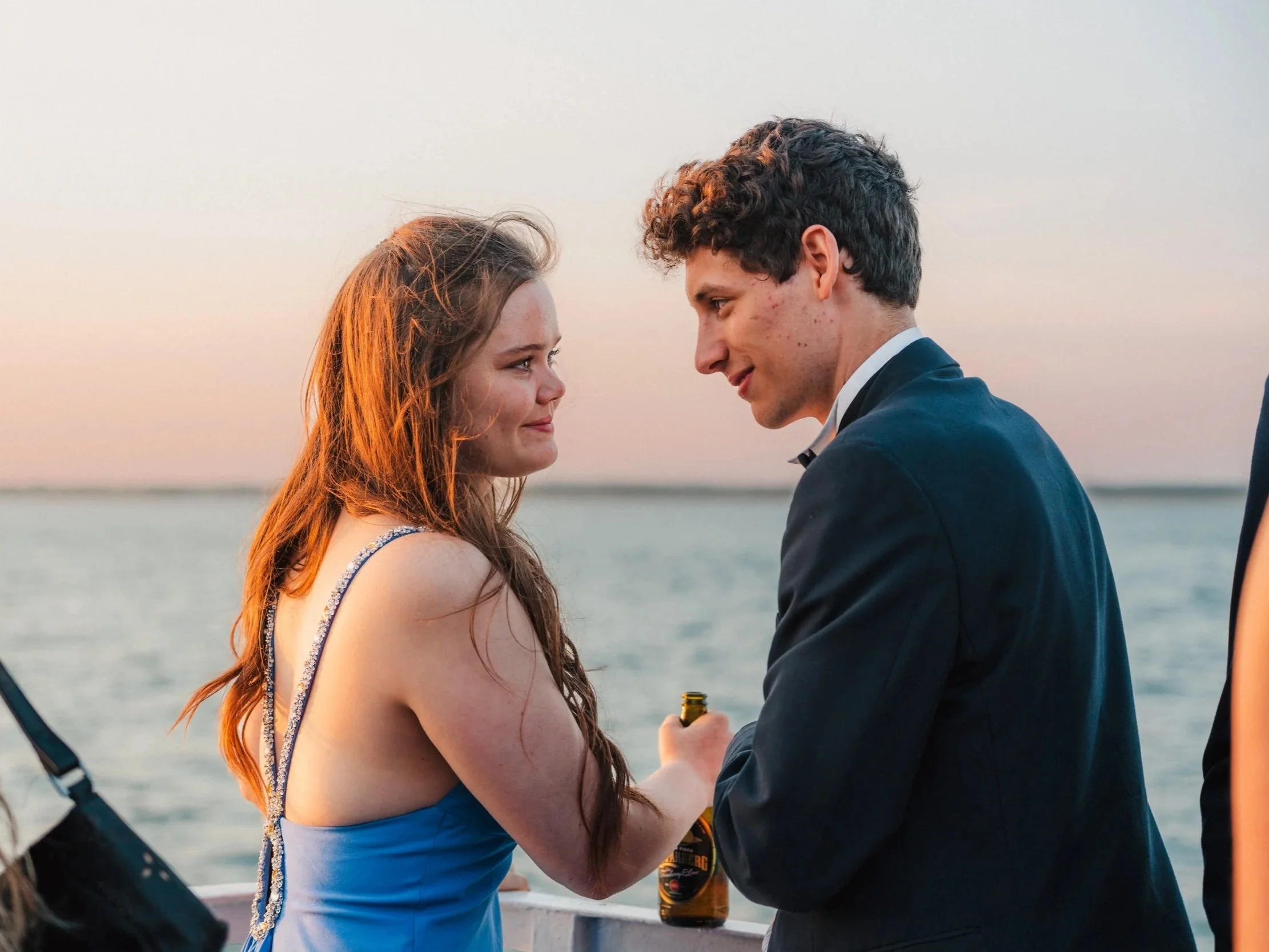 A young woman in a blue dress and a young man in a tuxedo having a romantic moment by the water at sunset, holding a beer bottle.