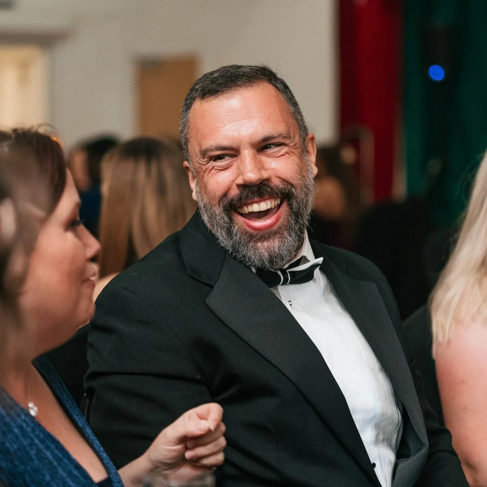 A man in a tuxedo with a bowtie, smiling and laughing at a formal event or party.