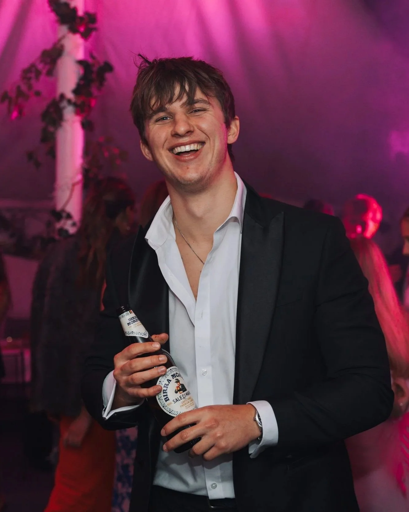 A young man in a tuxedo holding a beer bottle, smiling at a party with purple and pink lighting, with other people in the background.