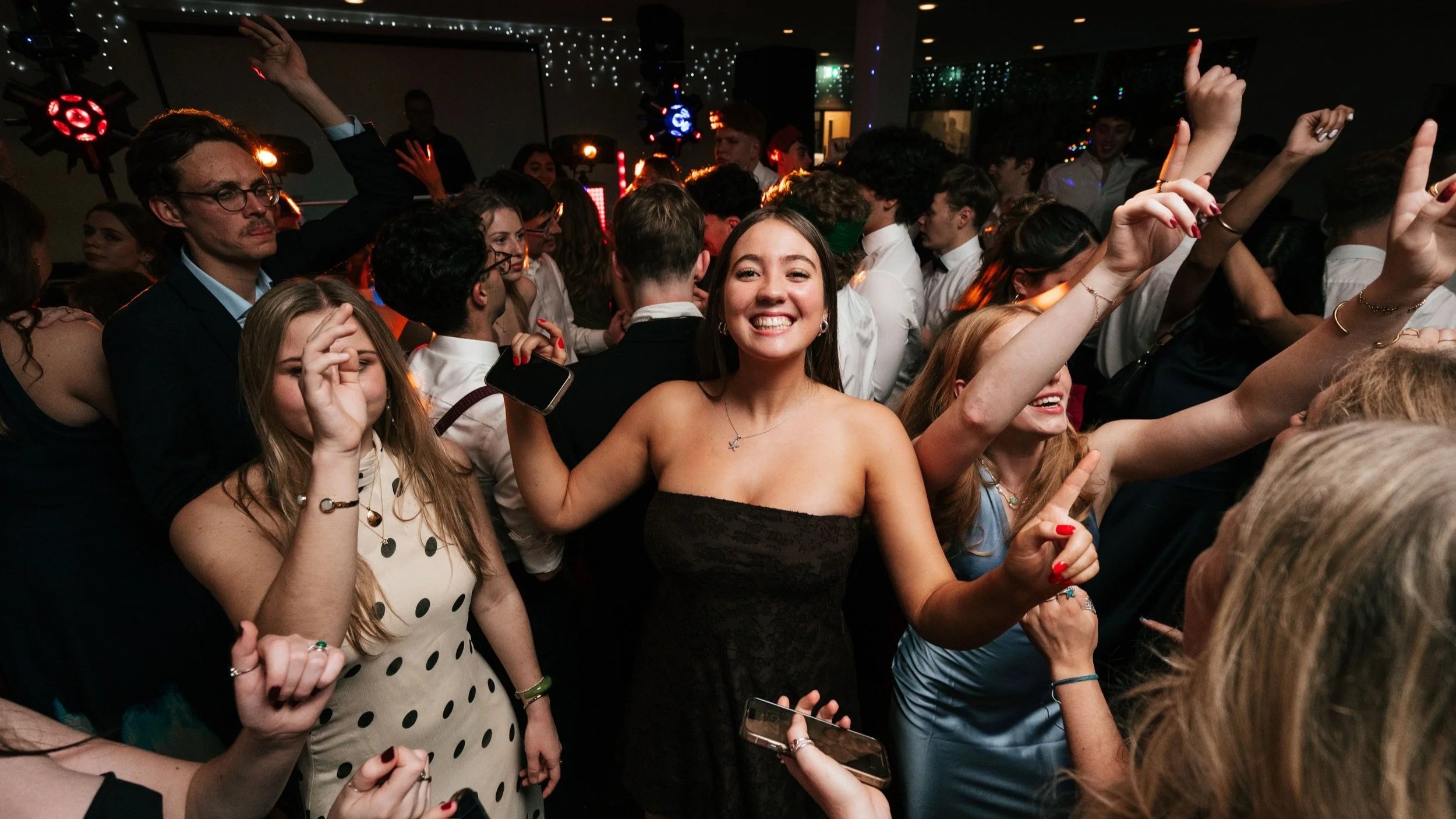 People dancing and celebrating at a lively party or dance floor, with smiles and raised hands, under colorful lighting.