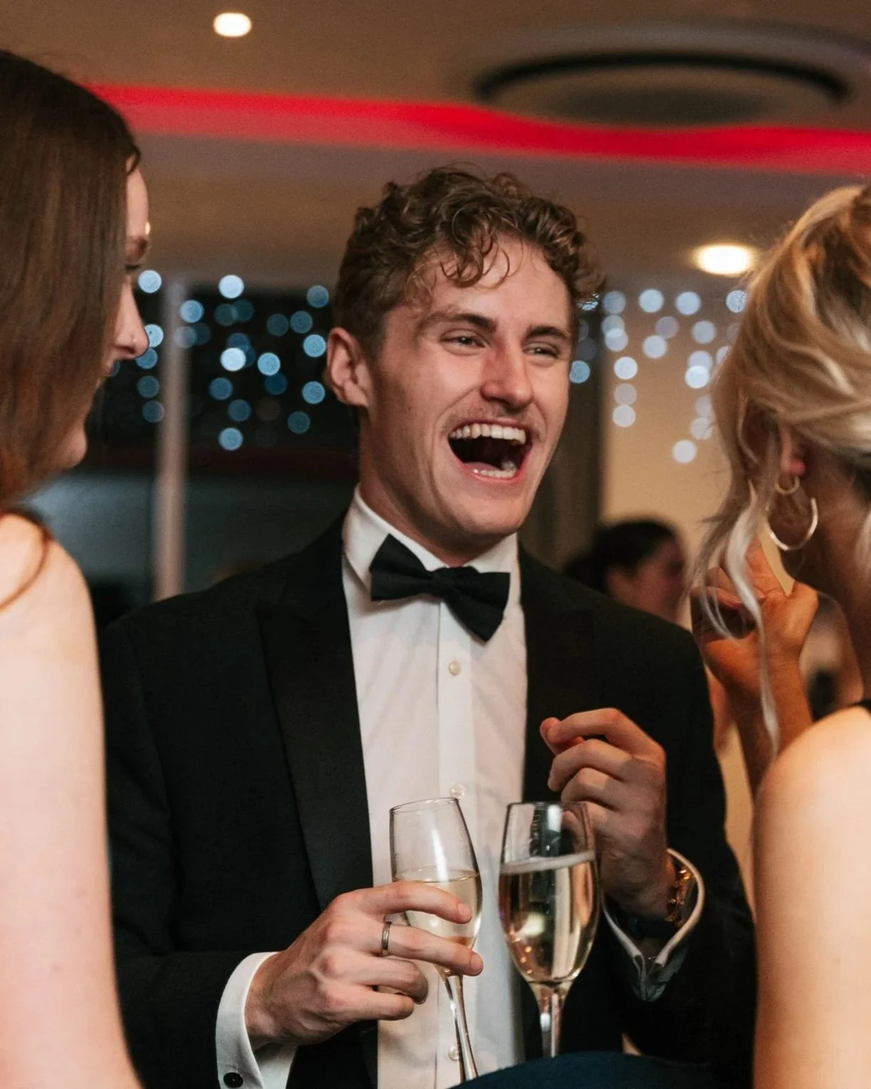 Young man in tuxedo with bow tie smiling and laughing at a party, holding a glass of champagne, surrounded by women.