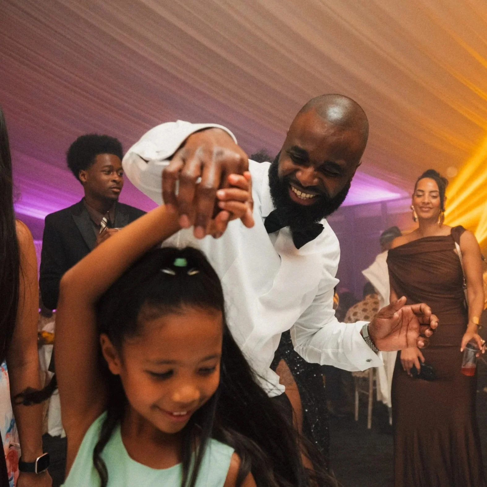 A man with a beard dancing with a young girl at a party. The man is smiling and wearing a white shirt with a black bow tie. The girl is smiling and has her arm raised. Other adults are dancing and socializing in the background, with colorful lighting