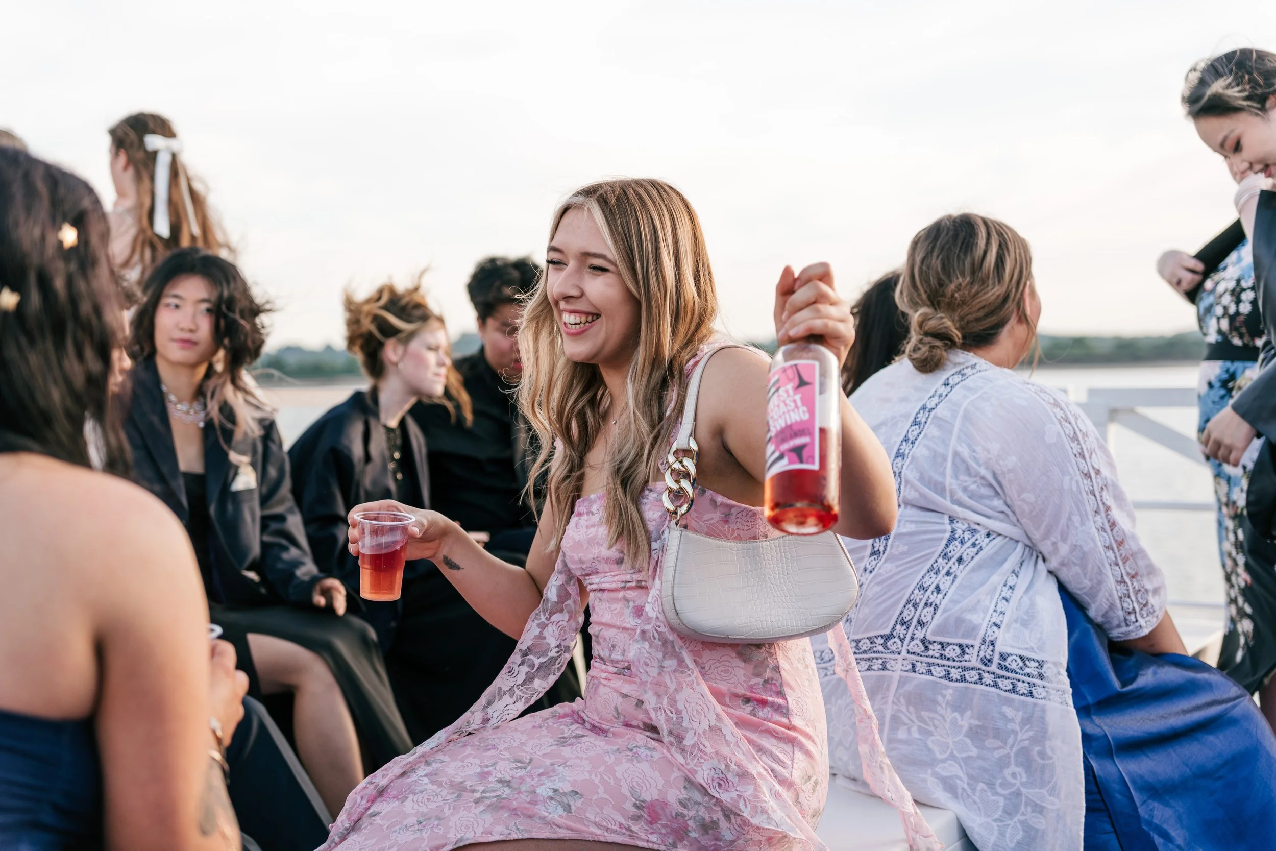 A smiling woman in a pink dress holding a bottle and a cup of drinks at a gathering by the water with other people sitting nearby.