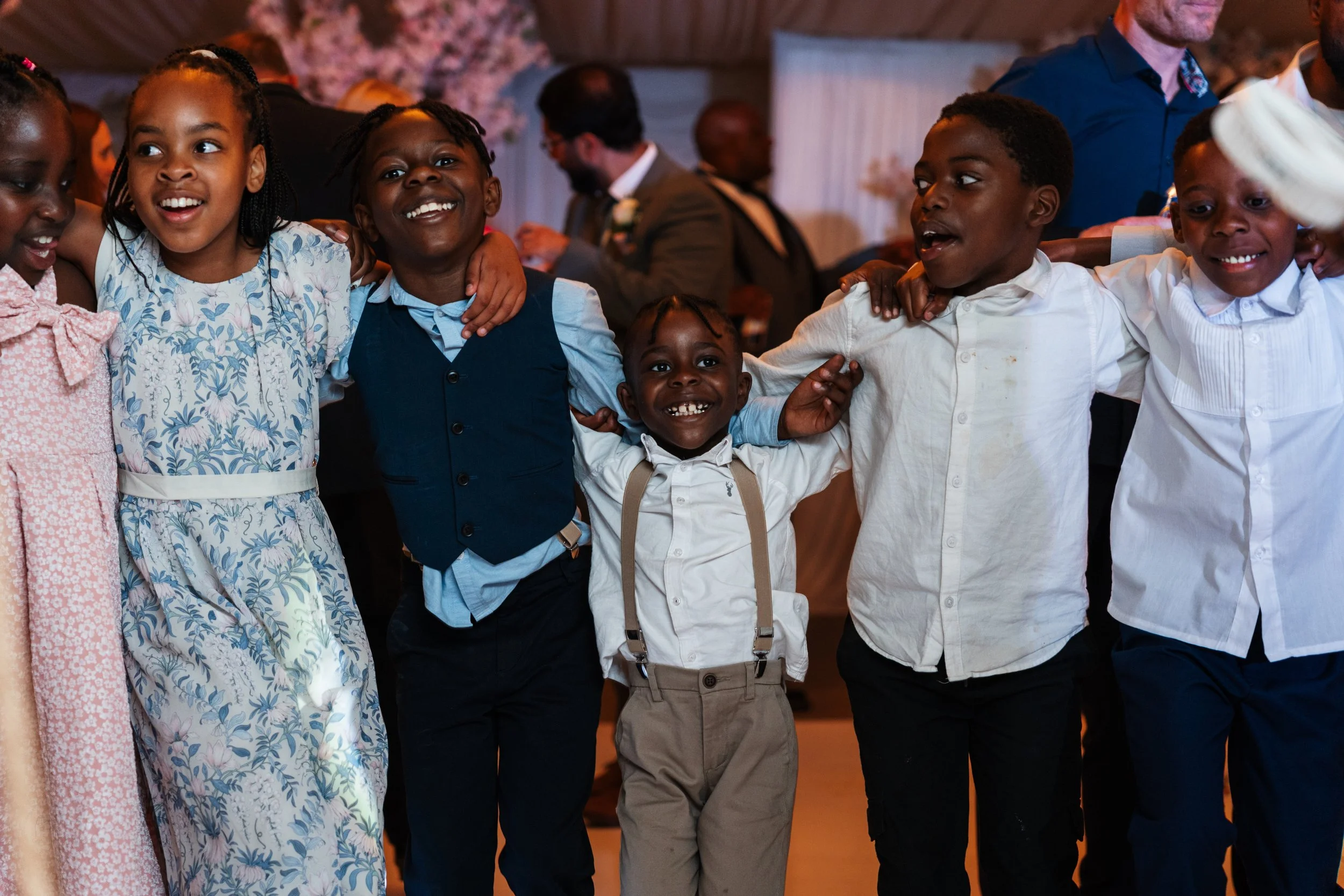 A group of seven children standing in a line with their arms around each other's shoulders at a celebration or party, smiling and looking happy.