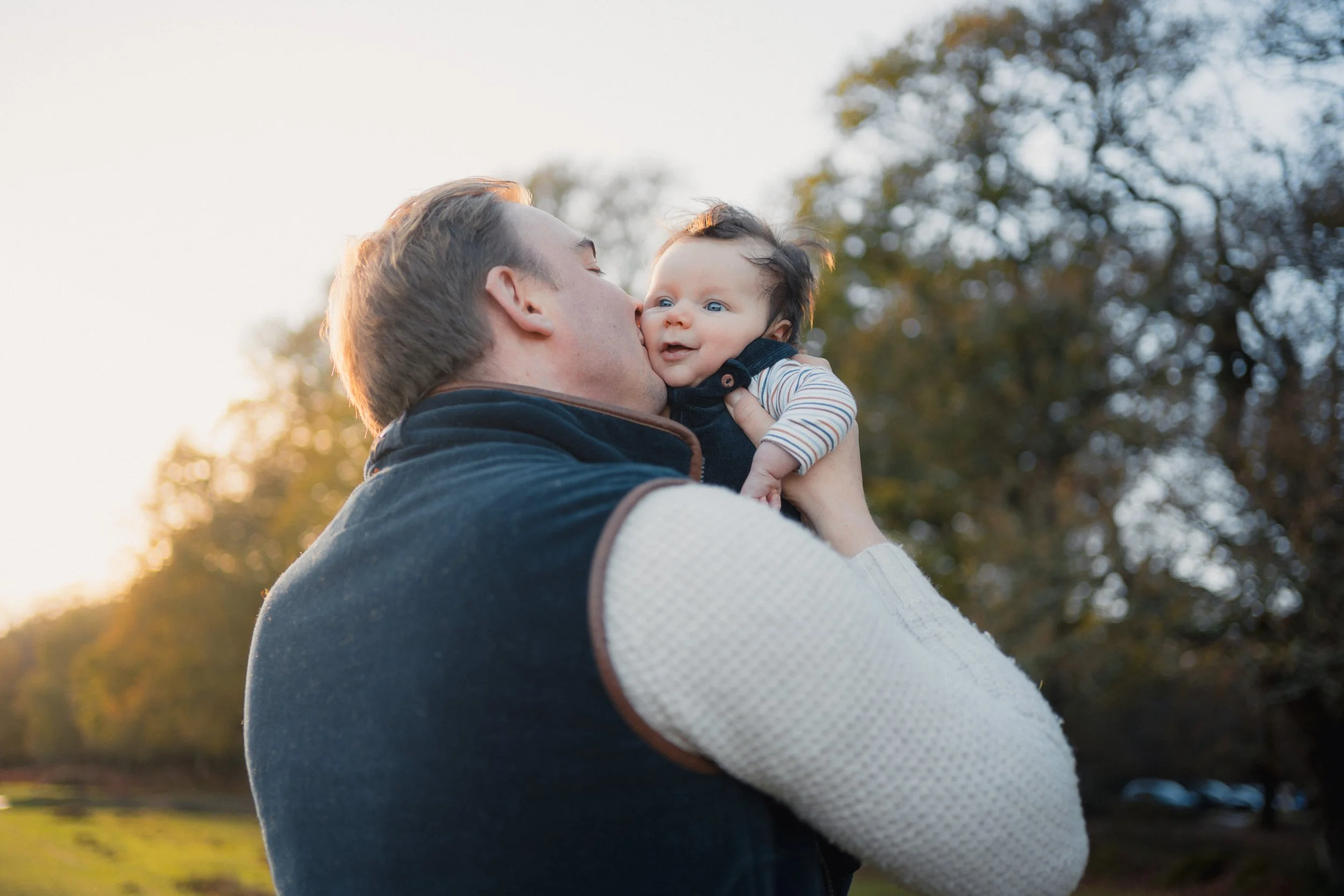 A man holding a baby outdoors at sunset, trees in the background.