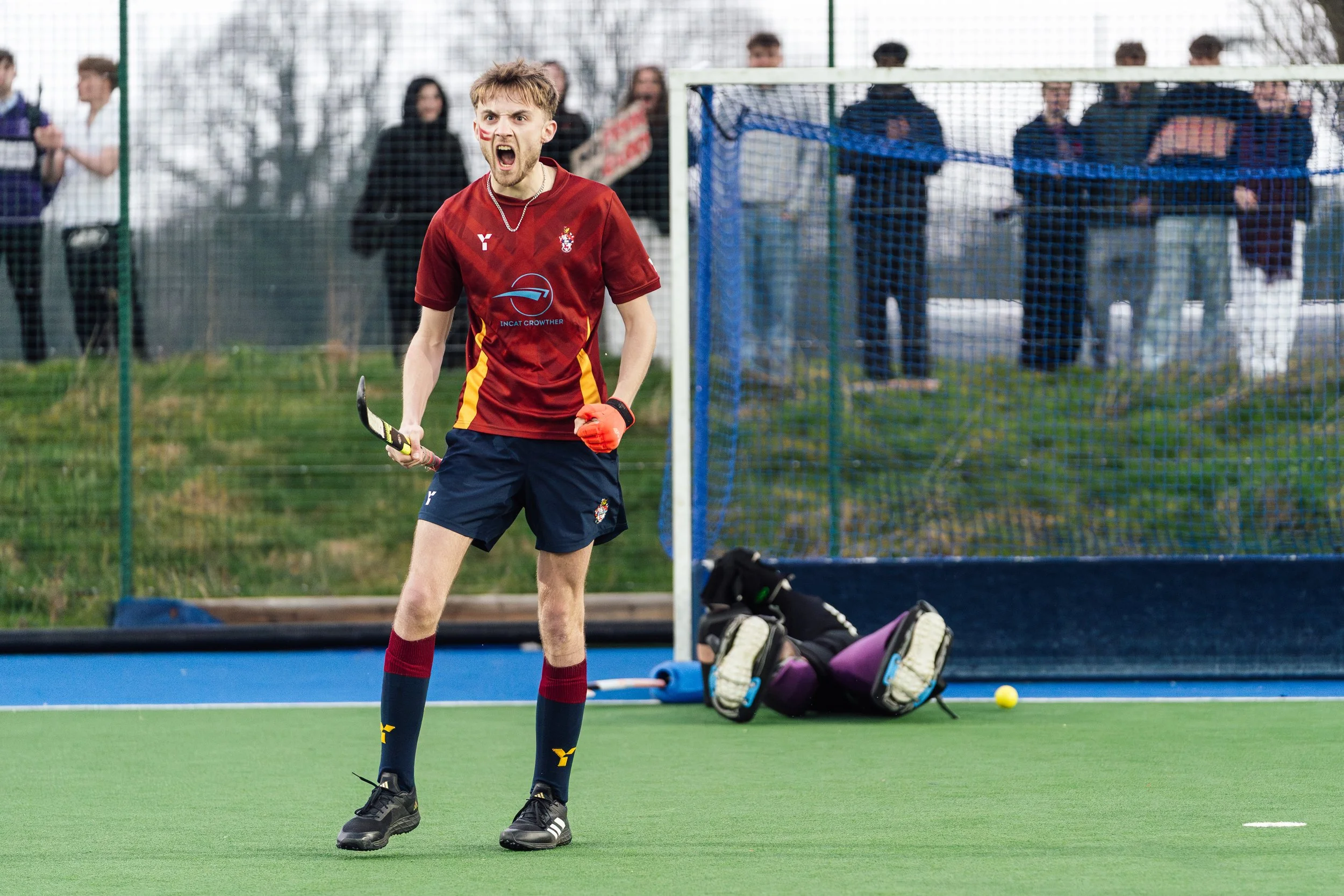 Field hockey goalie on the ground with a helmet and pads, while a player in a red and yellow uniform celebrates on the field, with spectators watching from behind a fence.