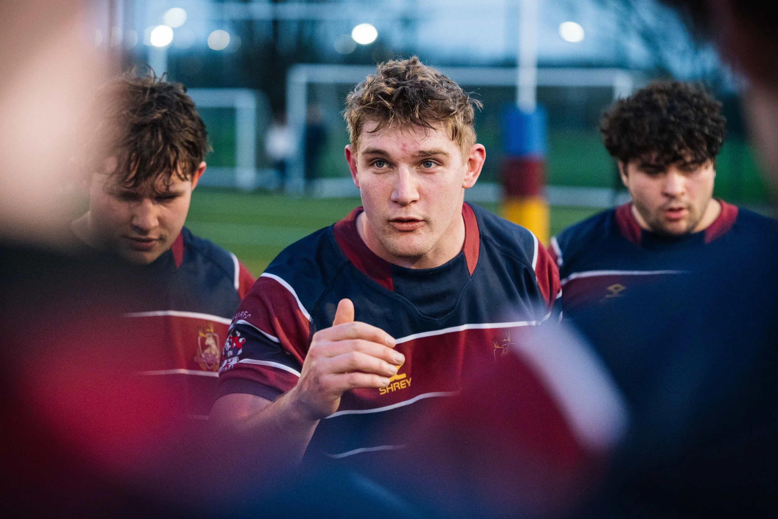 A young rugby player with short, light brown hair and a serious expression, wearing a navy blue and maroon rugby jersey, speaking to teammates during a game or practice on a grassy field.