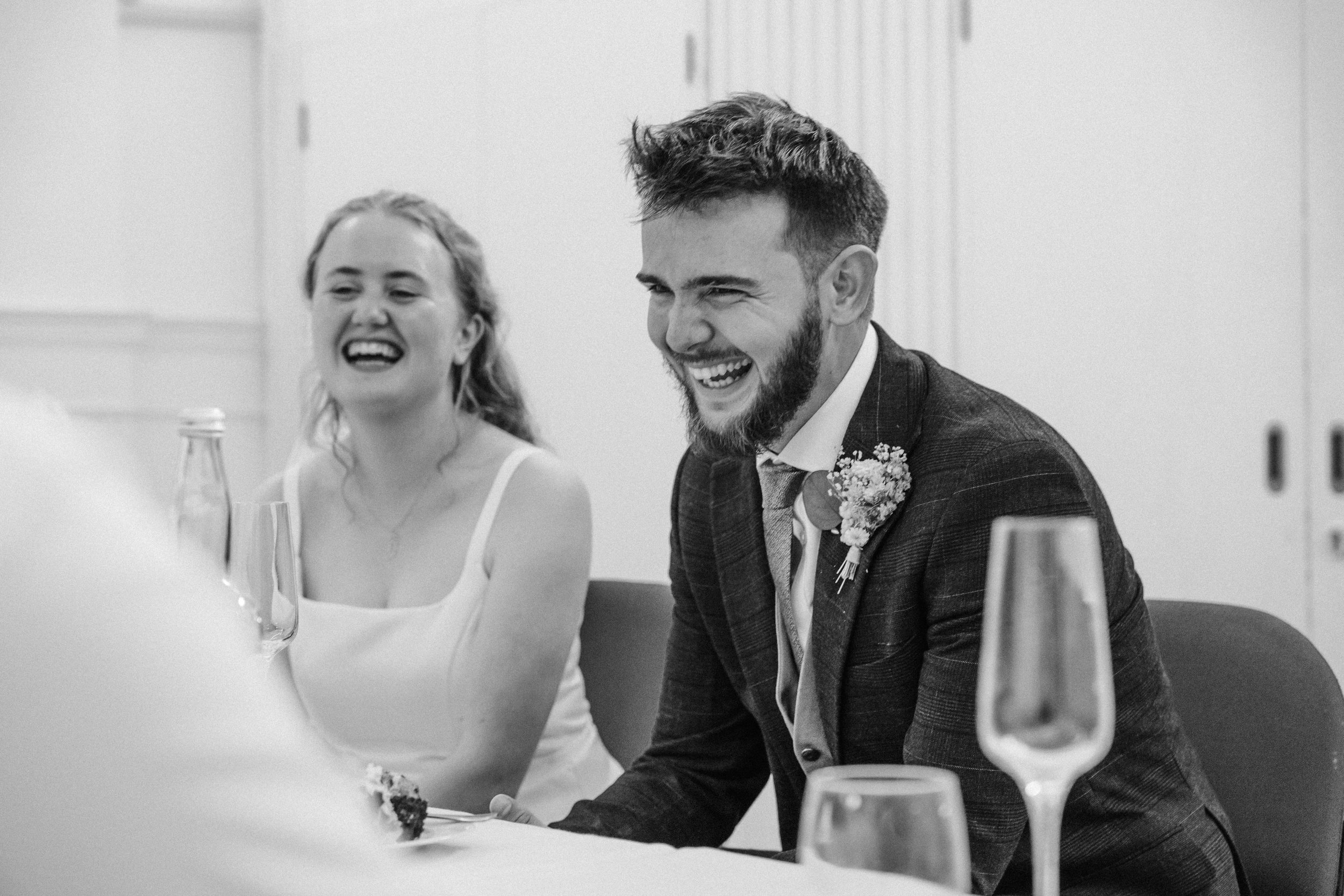 A man and woman laughing at a wedding reception. The woman is wearing a white dress, and the man is dressed in a suit with a boutonniere.