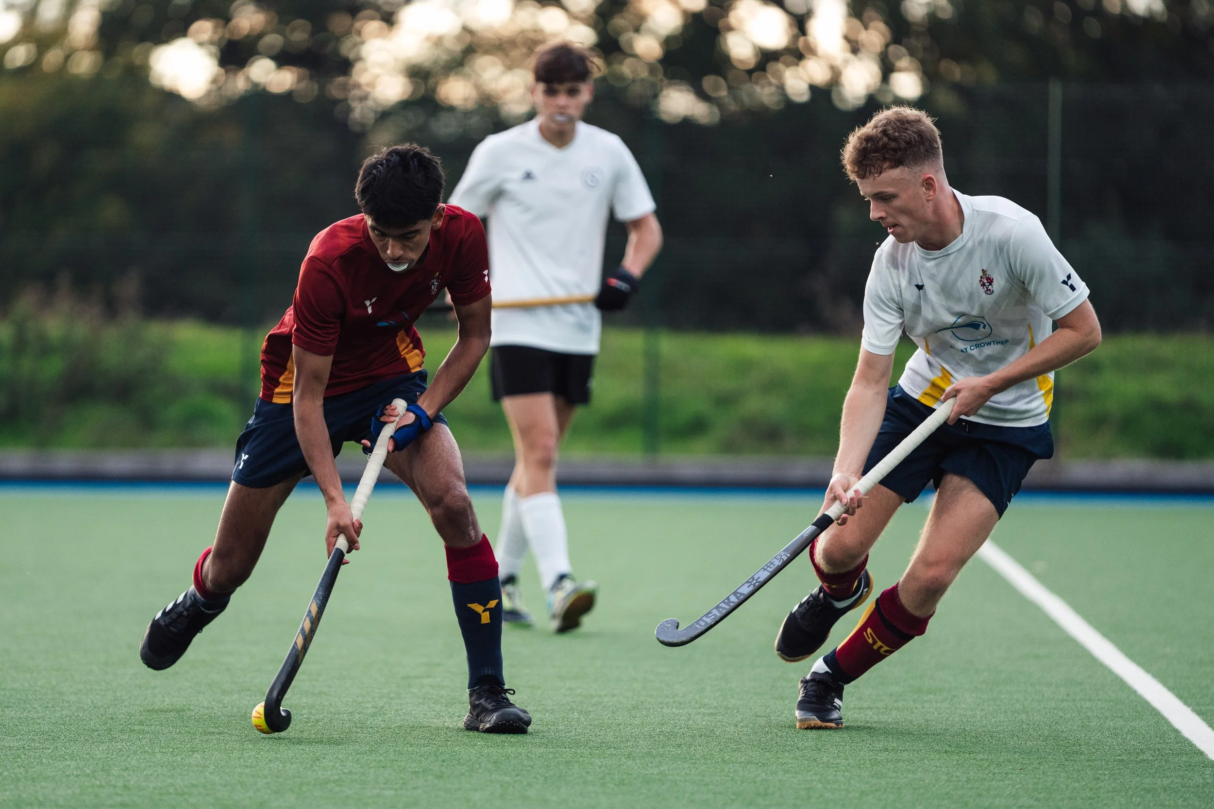 Two young men playing field hockey, one in a maroon jersey and the other in a white jersey, on a green sports field with a third person in the background.
