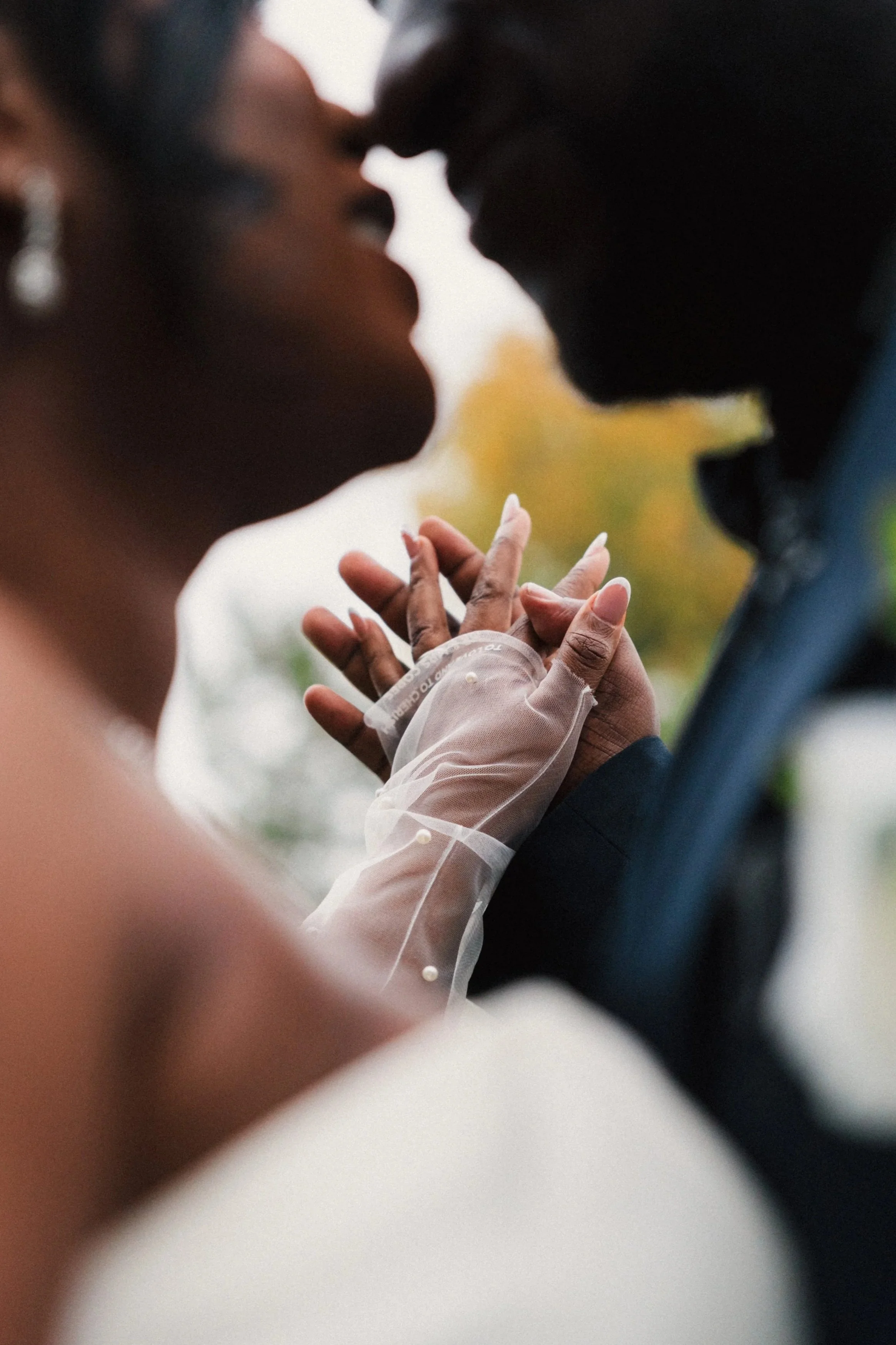 Close-up of a couple about to kiss, holding hands, with one person wearing a sheer, long-sleeved dress with pearl accents, outdoors with blurred background.