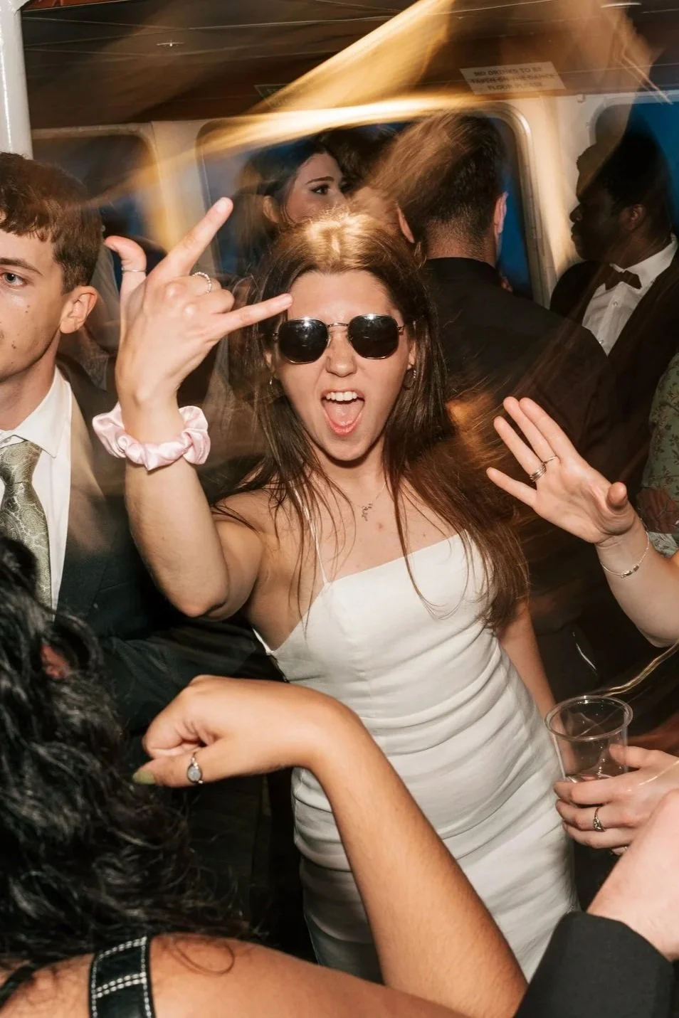 A young woman with dark hair, wearing sunglasses and a white dress, making a rock-and-roll hand gesture at a party or celebration.