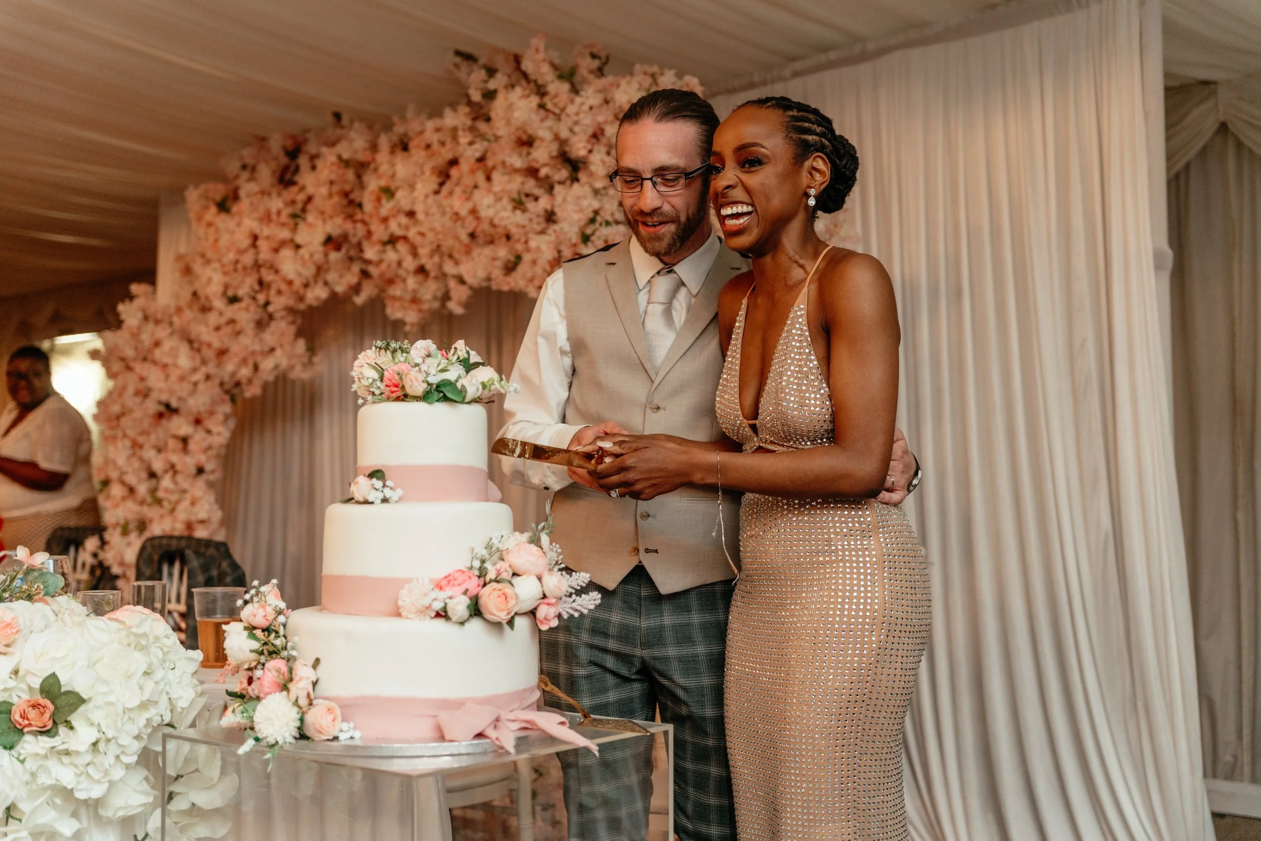 A bride and groom cutting a wedding cake together at their wedding reception, decorated with pink and white flowers and a pink ribbon, with a floral arch in the background.