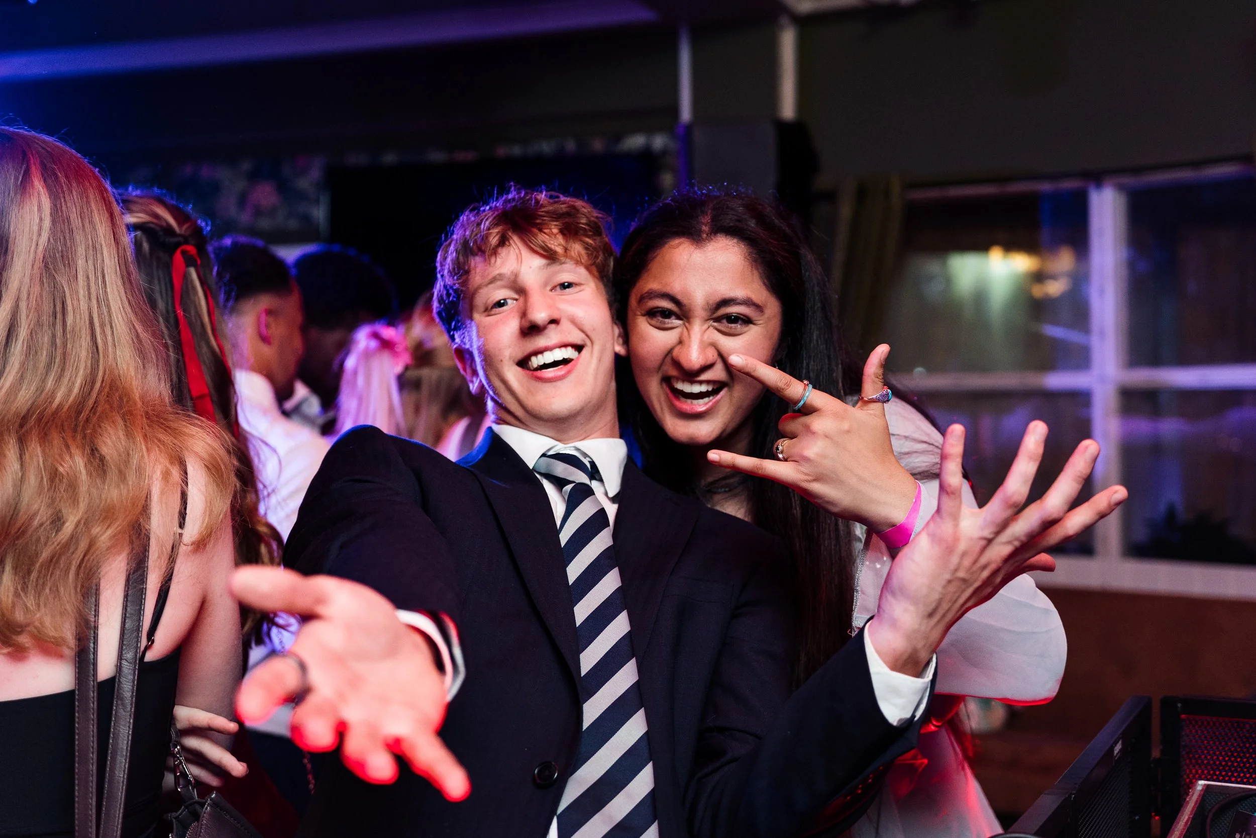 Two young people at a party or celebration, smiling and having fun; one with orange hair in a suit and striped tie, the other with dark hair making a rock-and-roll gesture.