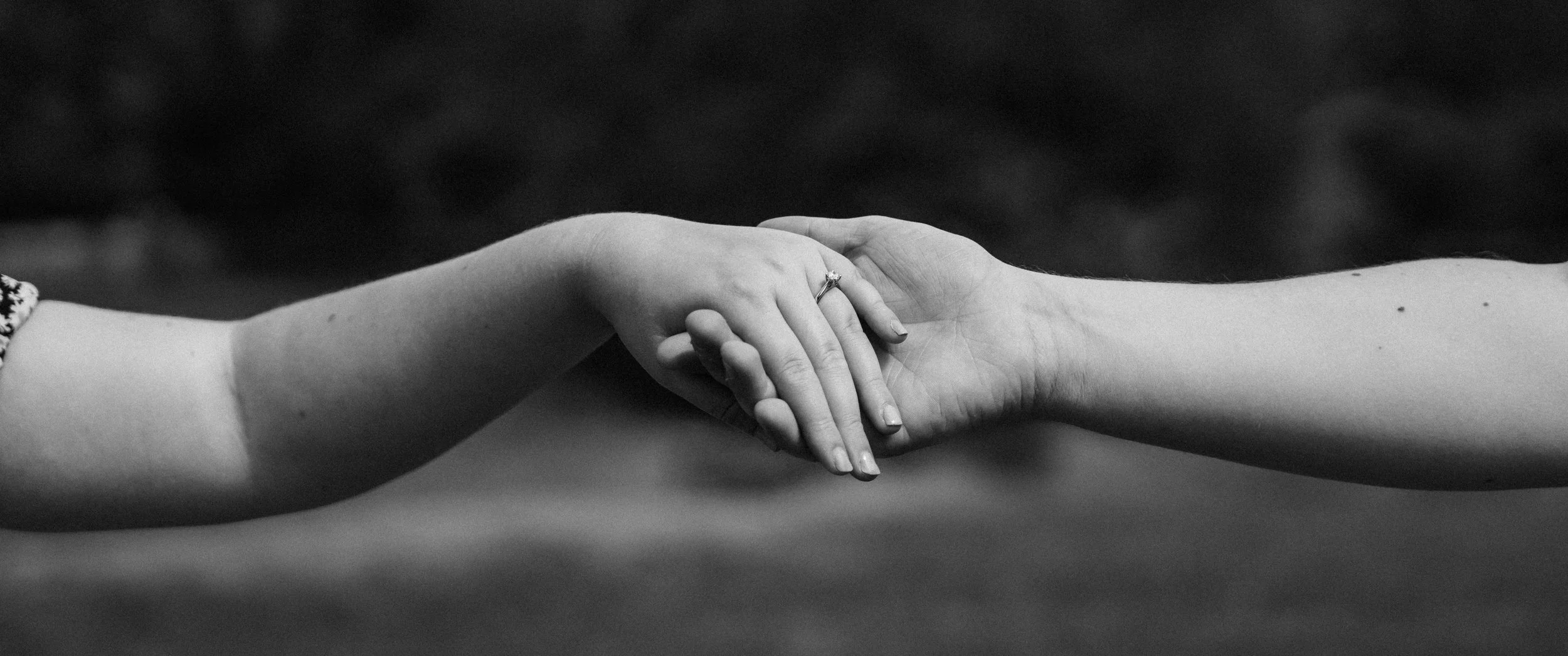 A close-up black and white photograph of two hands holding each other, one child and one adult, with a dark blurred background.