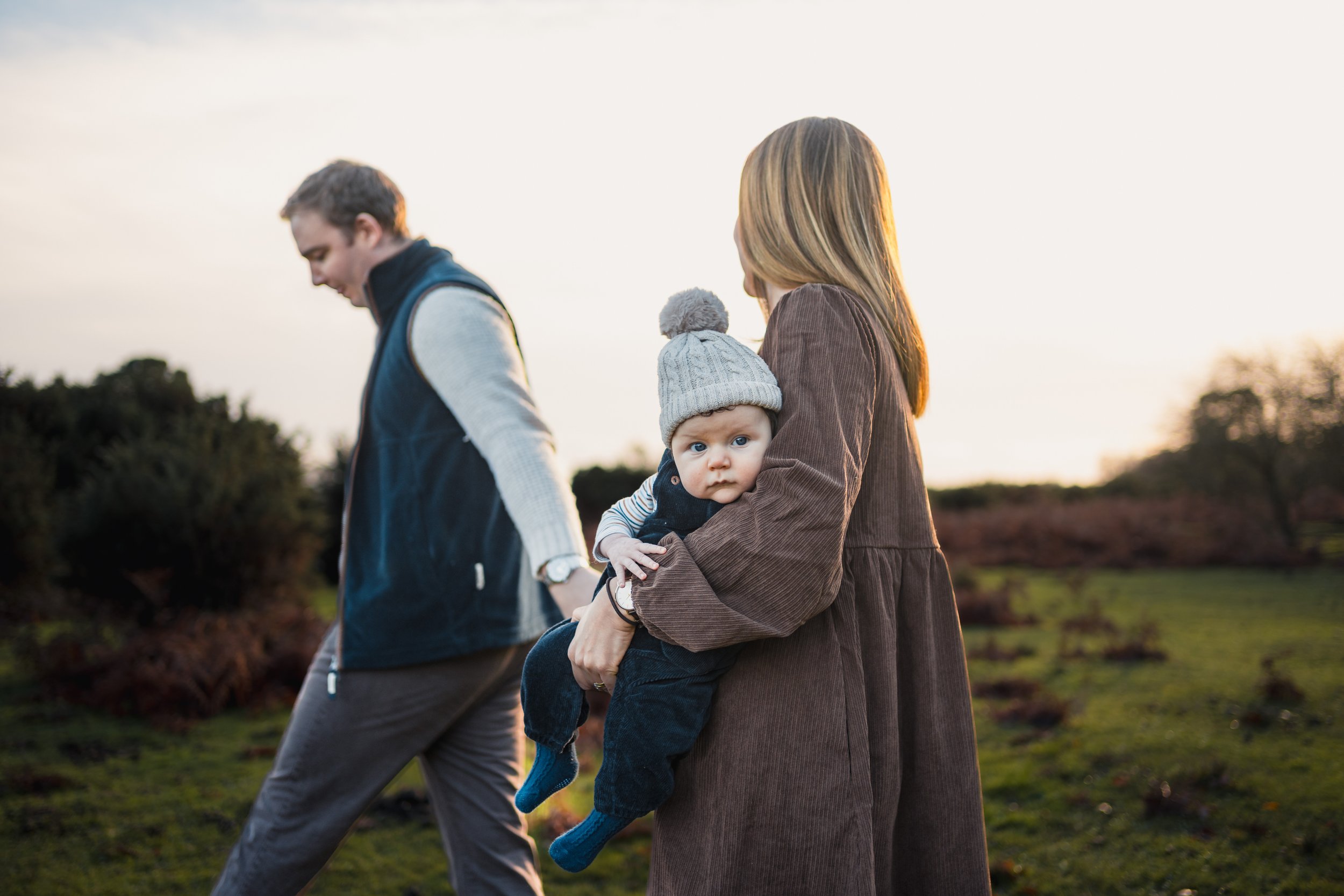 A woman holding a baby with a knit hat, walking outdoors at sunset, with a man walking ahead in the background.