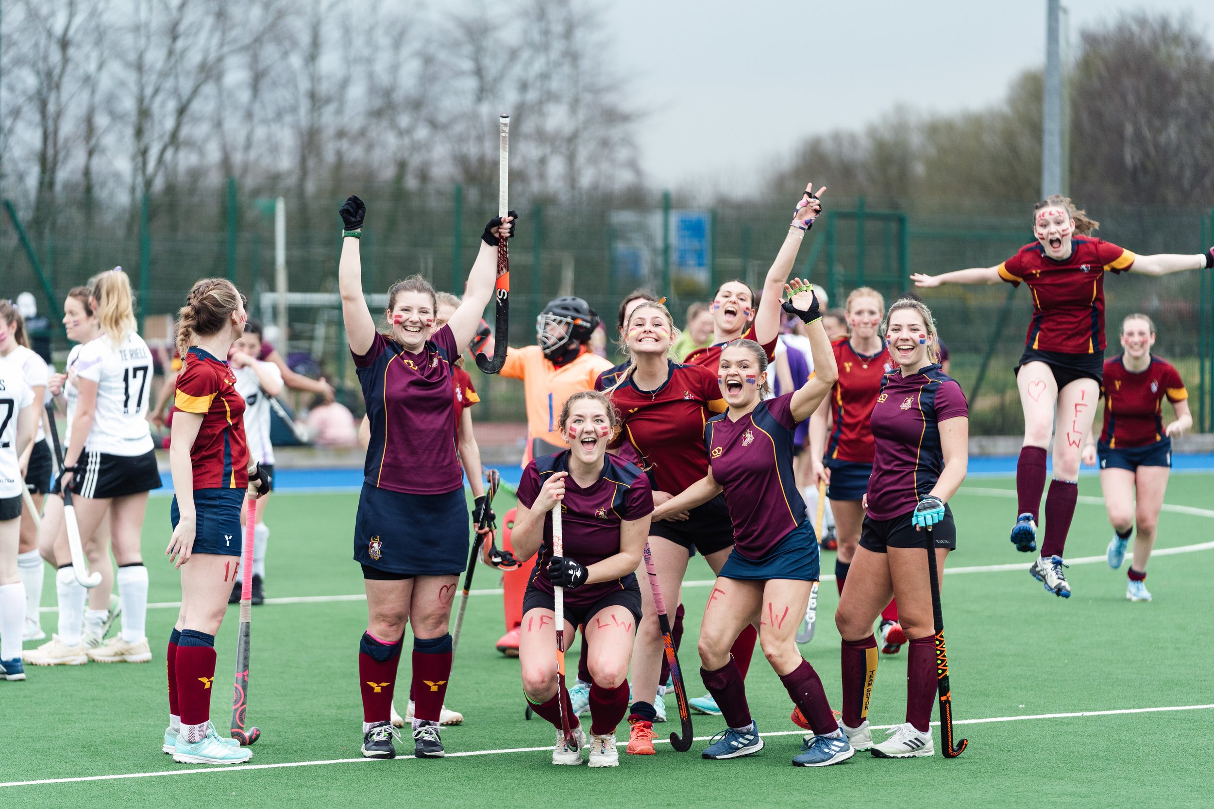 Women’s field hockey team celebrating on the field, some jumping and smiling with their sticks raised, in maroon and navy uniforms.