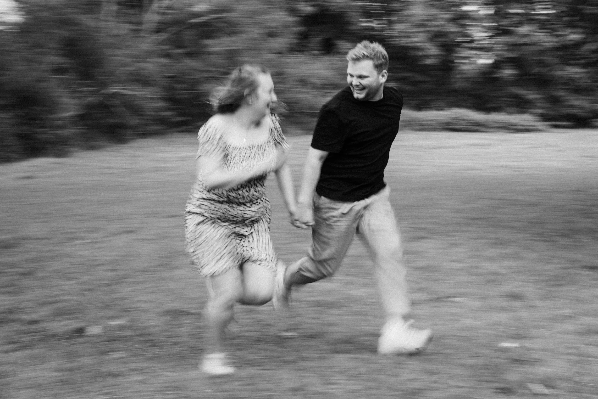A black and white photo of a man and woman running and holding hands outdoors, with trees in the background.