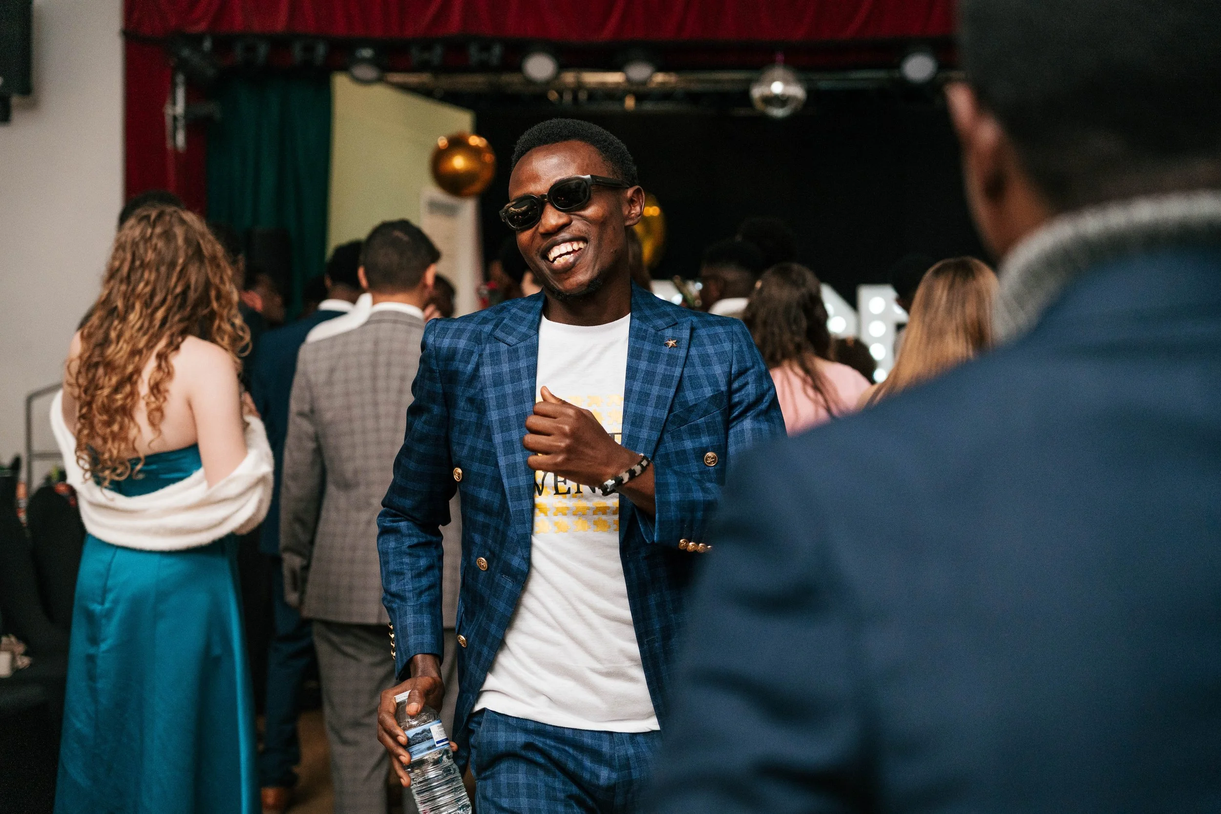 A man wearing sunglasses and a blue checkered suit jacket smiling and holding a water bottle, at a social event with several people in the background.