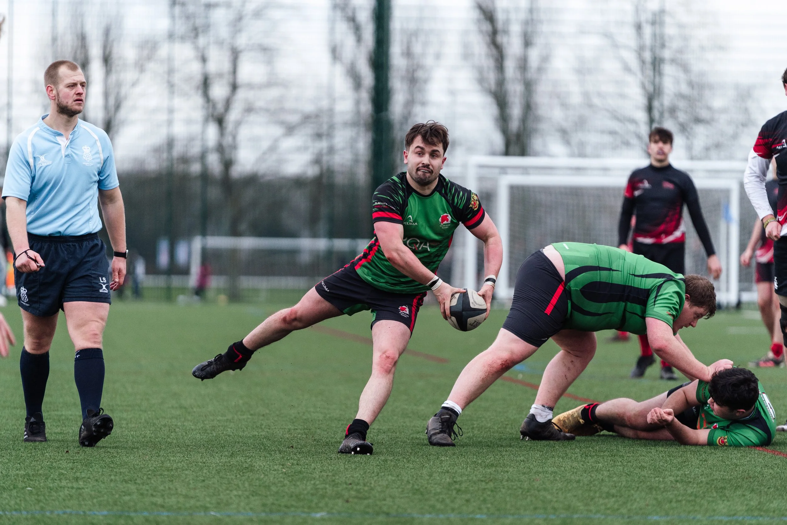 Rugby players in green and black uniforms compete for the ball during a game, with one player holding the ball, another on the ground, and a referee observing on the field.