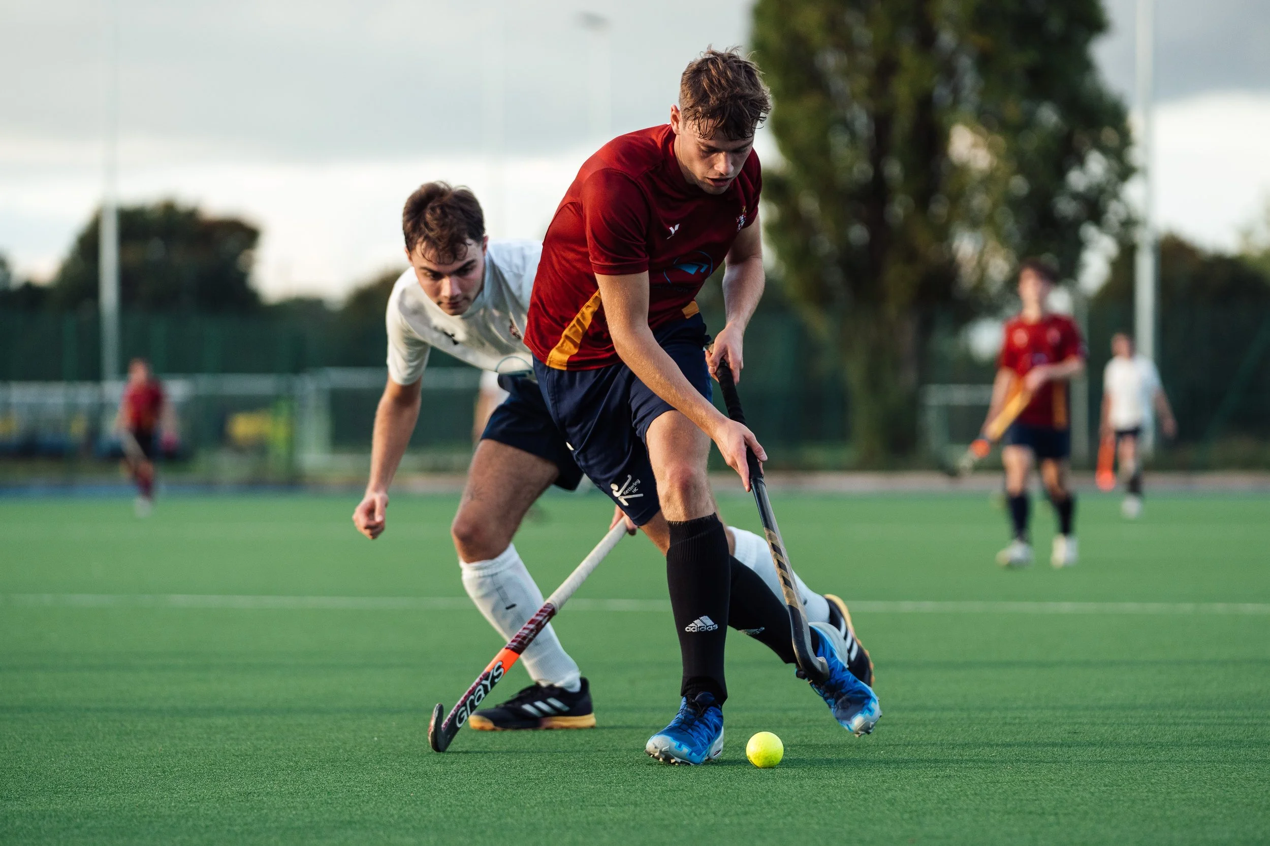 Two young male field hockey players in action on a green turf, one in a maroon and navy uniform, the other in a white and navy uniform, competing for the yellow ball during a game.