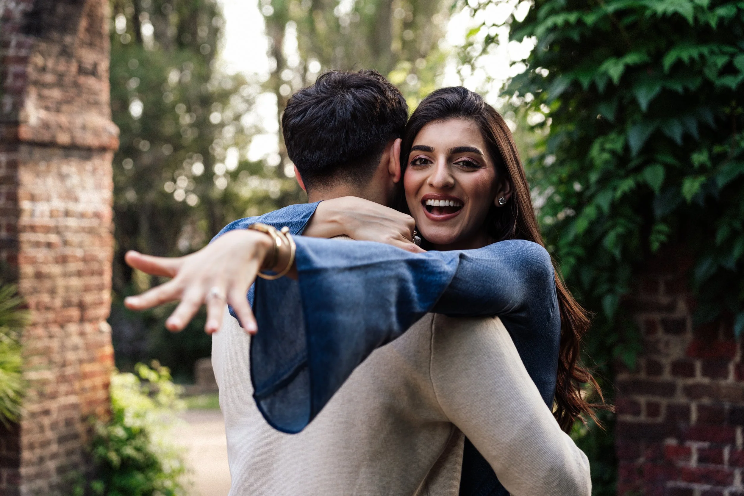 A smiling woman with long brown hair hugging a man with short dark hair outdoors.