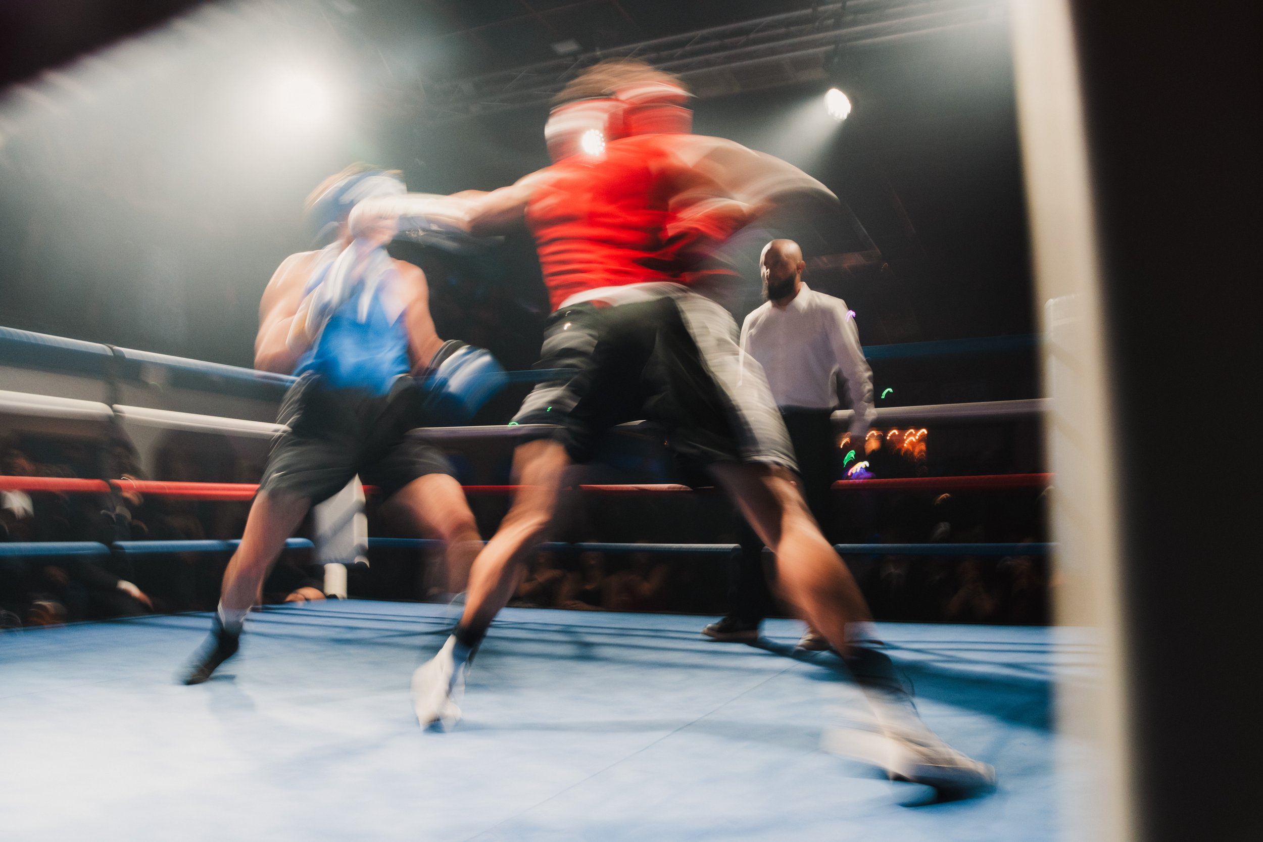 Two boxers in a boxing ring engaged in a fight, with a referee watching nearby and a blurred motion indicating fast movement.
