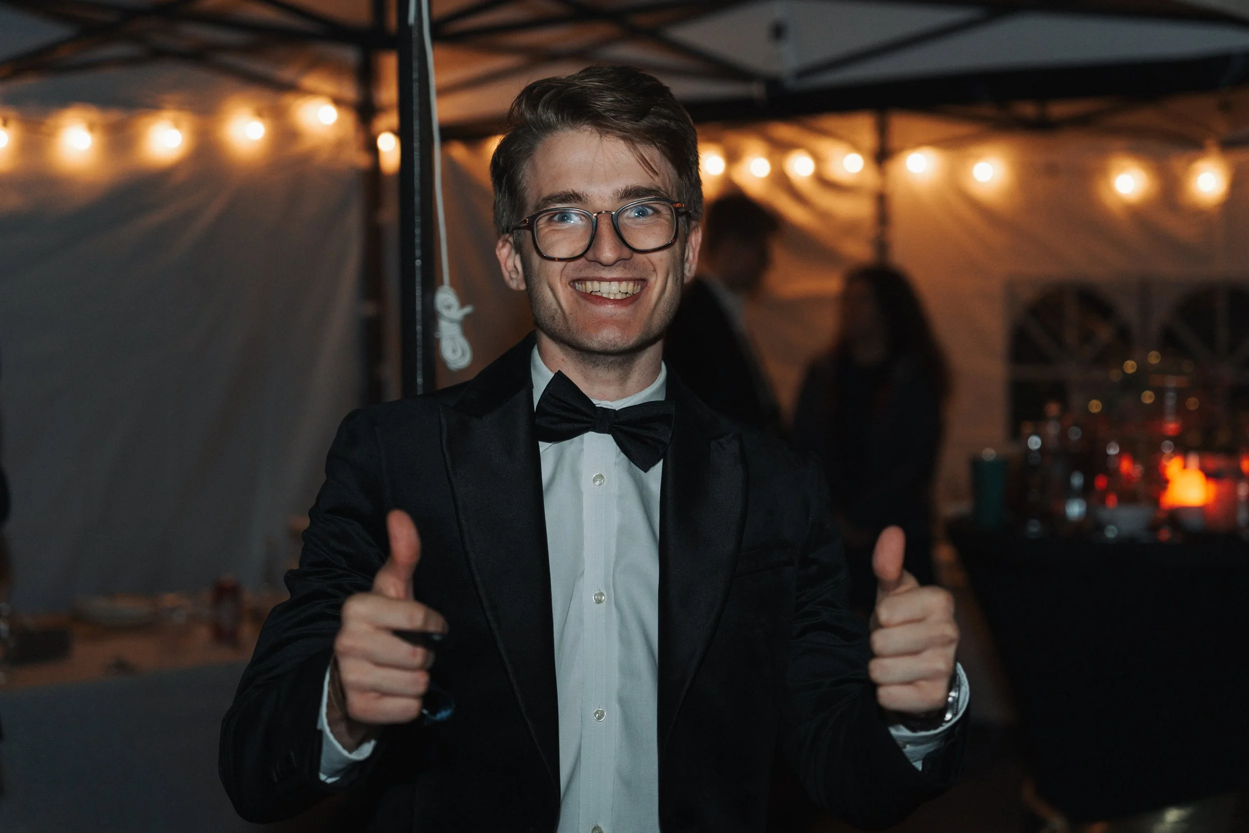 A smiling young man in a tuxedo and bow tie at a party or celebration, giving two thumbs up with string lights and a tent in the background.