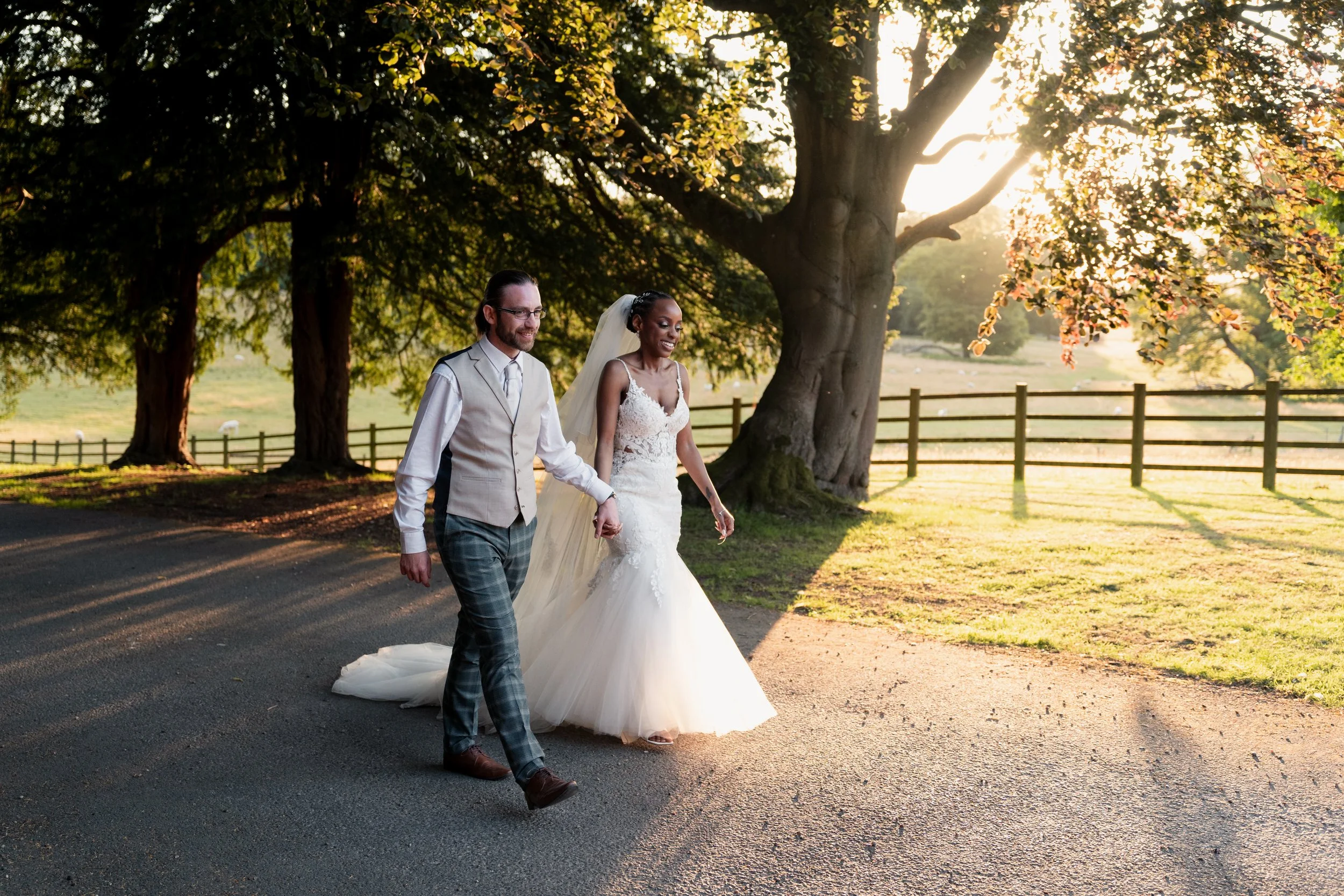A bride and groom walking hand-in-hand outdoors on a paved path during sunset, with a large tree and a wooden fence in the background.