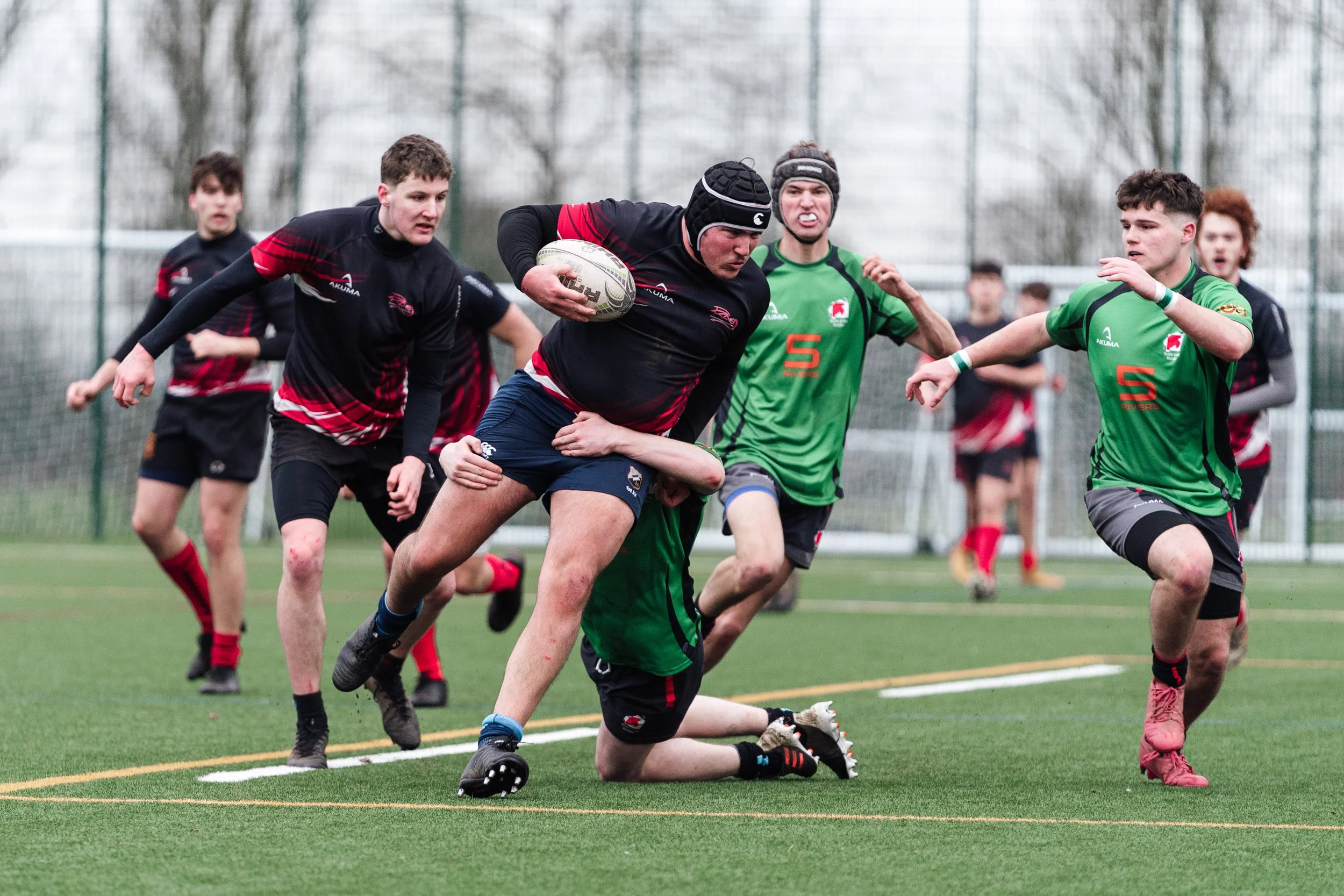 Rugby players in action on a field, with one player carrying the ball and others chasing him, during a match.
