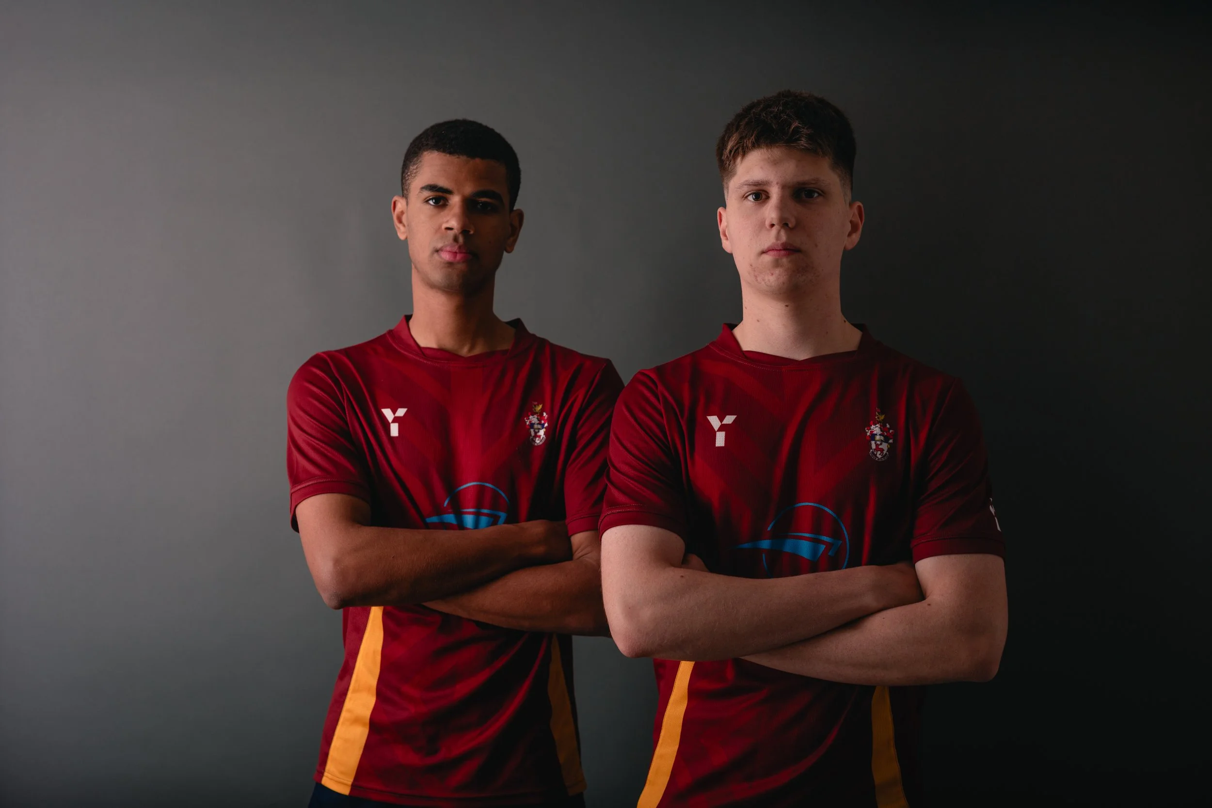 Two young men in maroon sports jerseys with yellow and black accents stand with arms crossed against a dark backdrop.