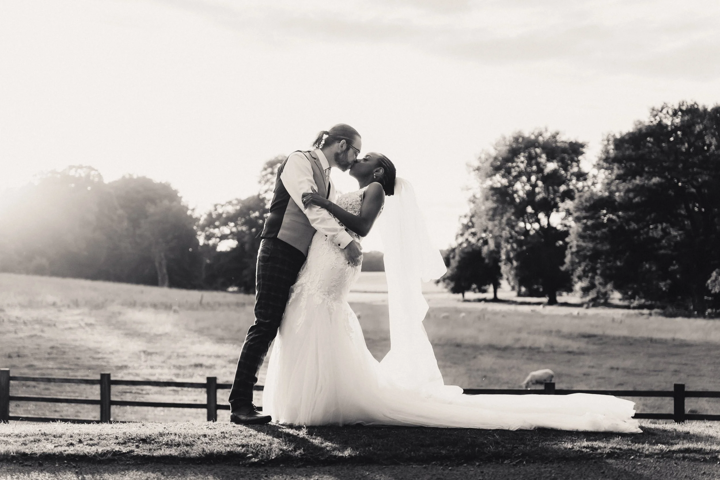 A black and white photo of a wedding couple kissing outdoors at sunset, with trees in the background and a fence in the foreground.