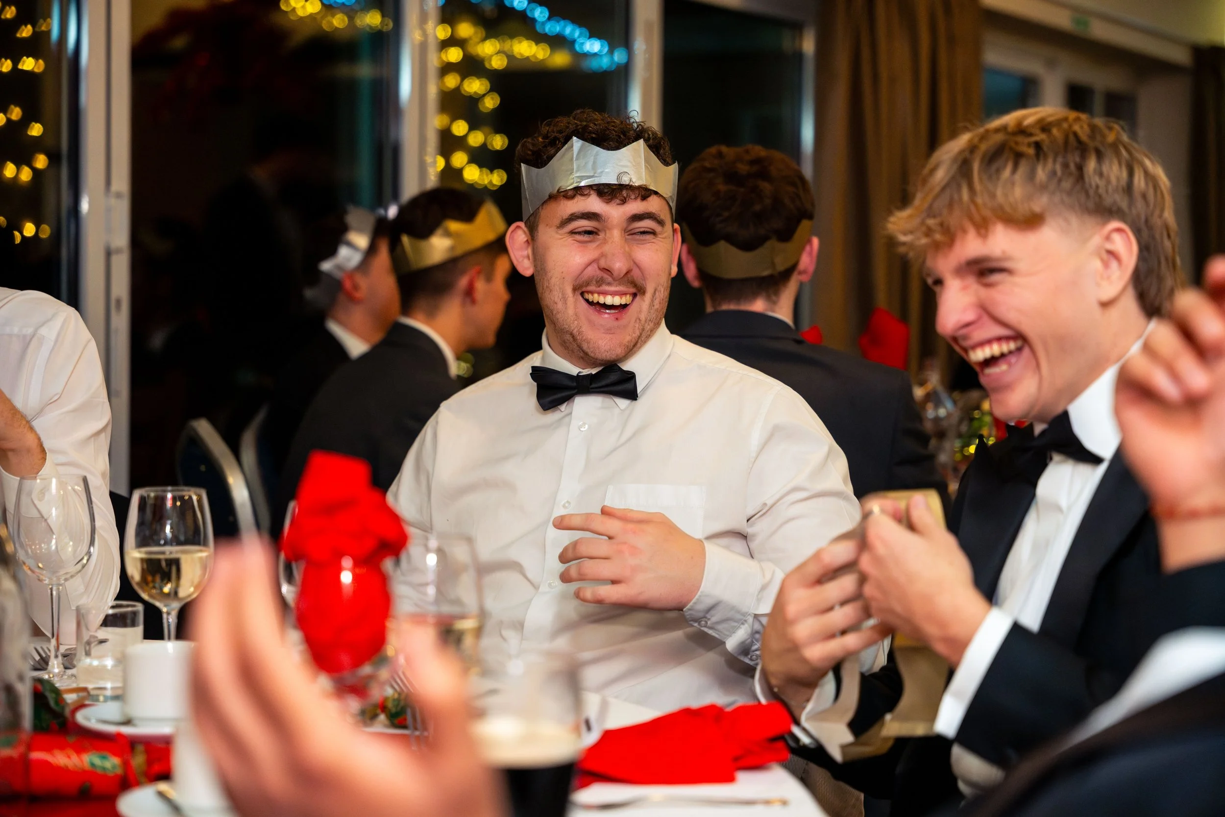 People dressed in tuxedos and white shirts wearing paper crowns, sitting at a table decorated for a celebration, smiling and laughing.