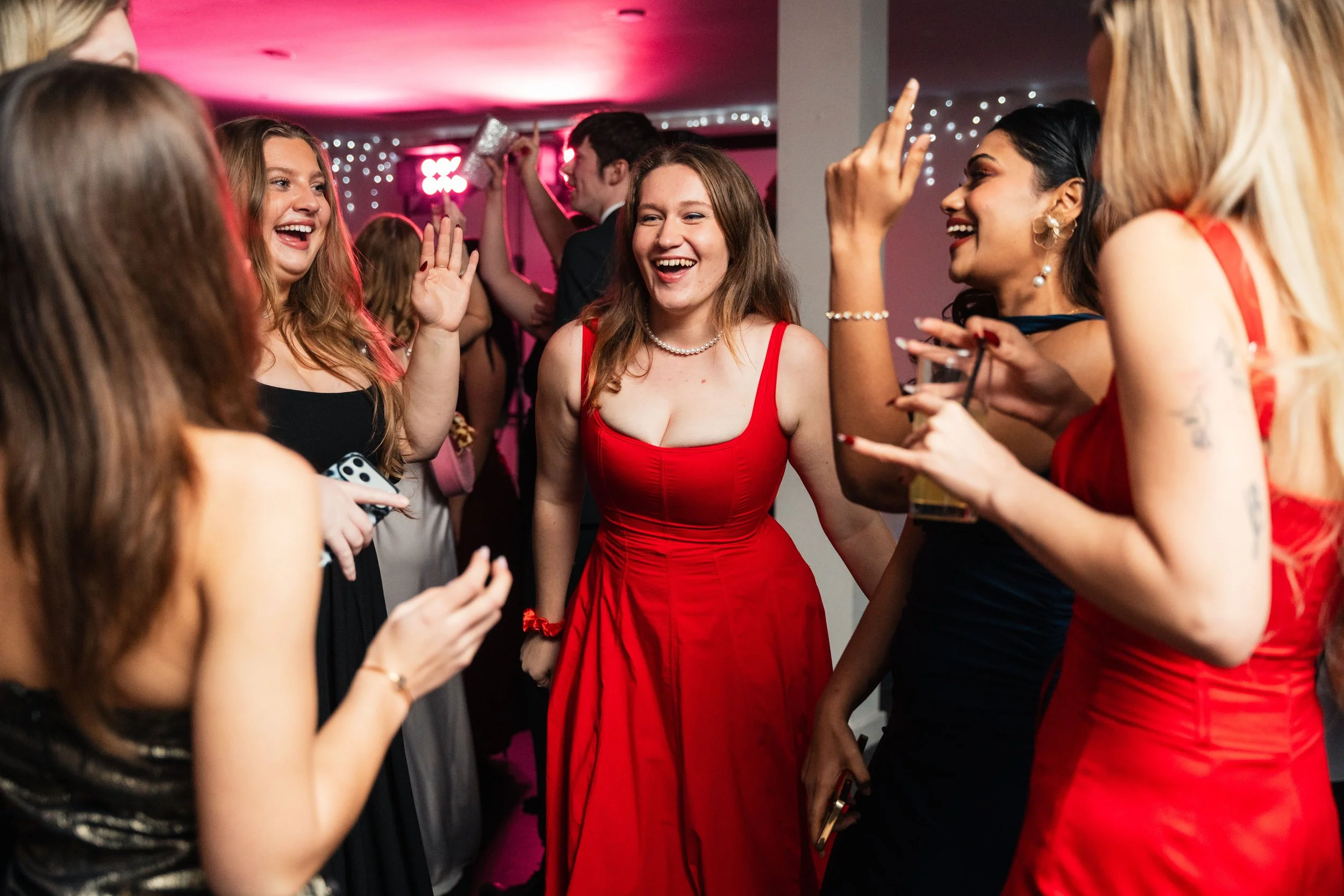 Group of women in evening dresses, laughing and talking at a lively party with pink and purple lighting and string lights in the background.