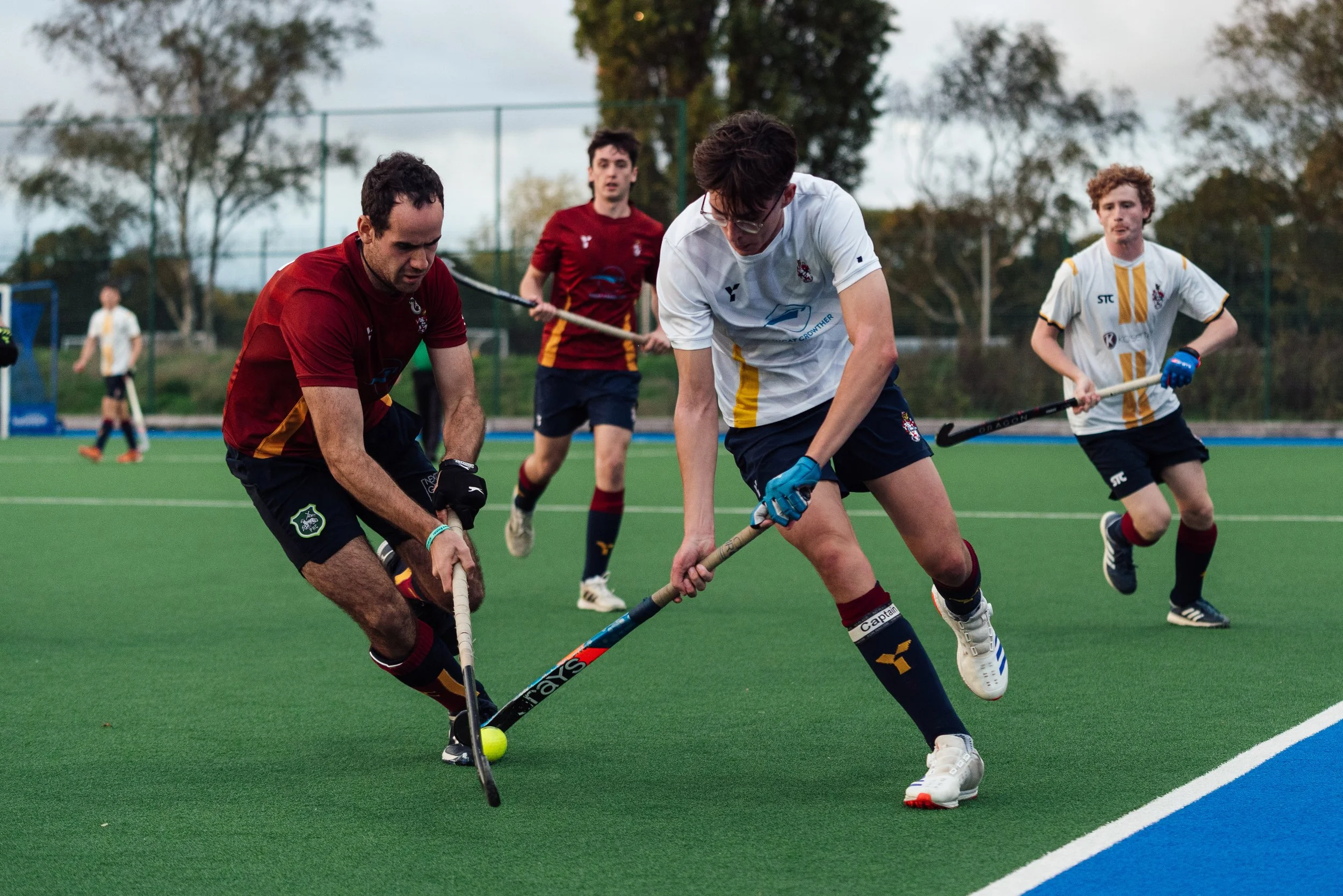 Two field hockey players competing for the ball on a green field, with other players in the background. One player in a white jersey and blue shorts, and the other in a maroon jersey and black shorts, are both bent over with their sticks near the yel