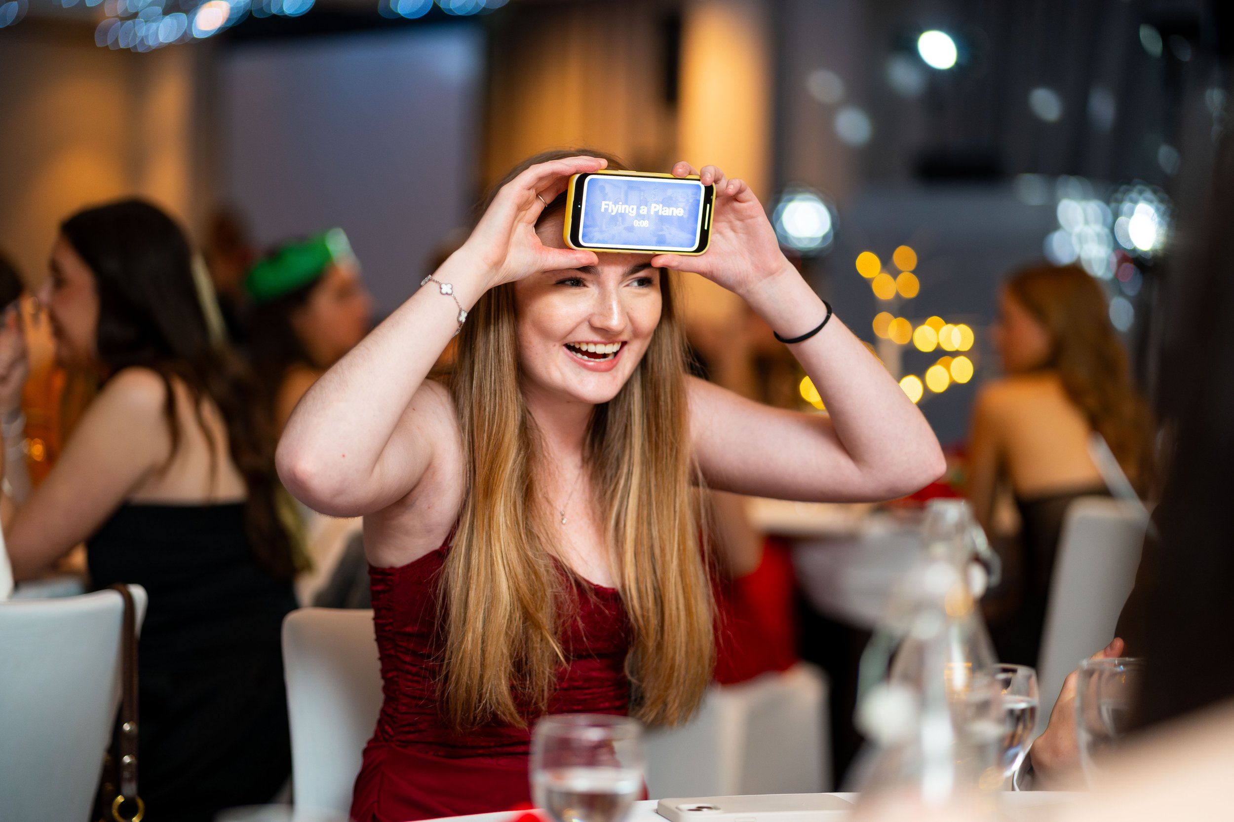 A woman with long red hair smiling and holding a phone above her head, with the screen displaying 'Flying a Plane,' at a party or celebration with others in the background.