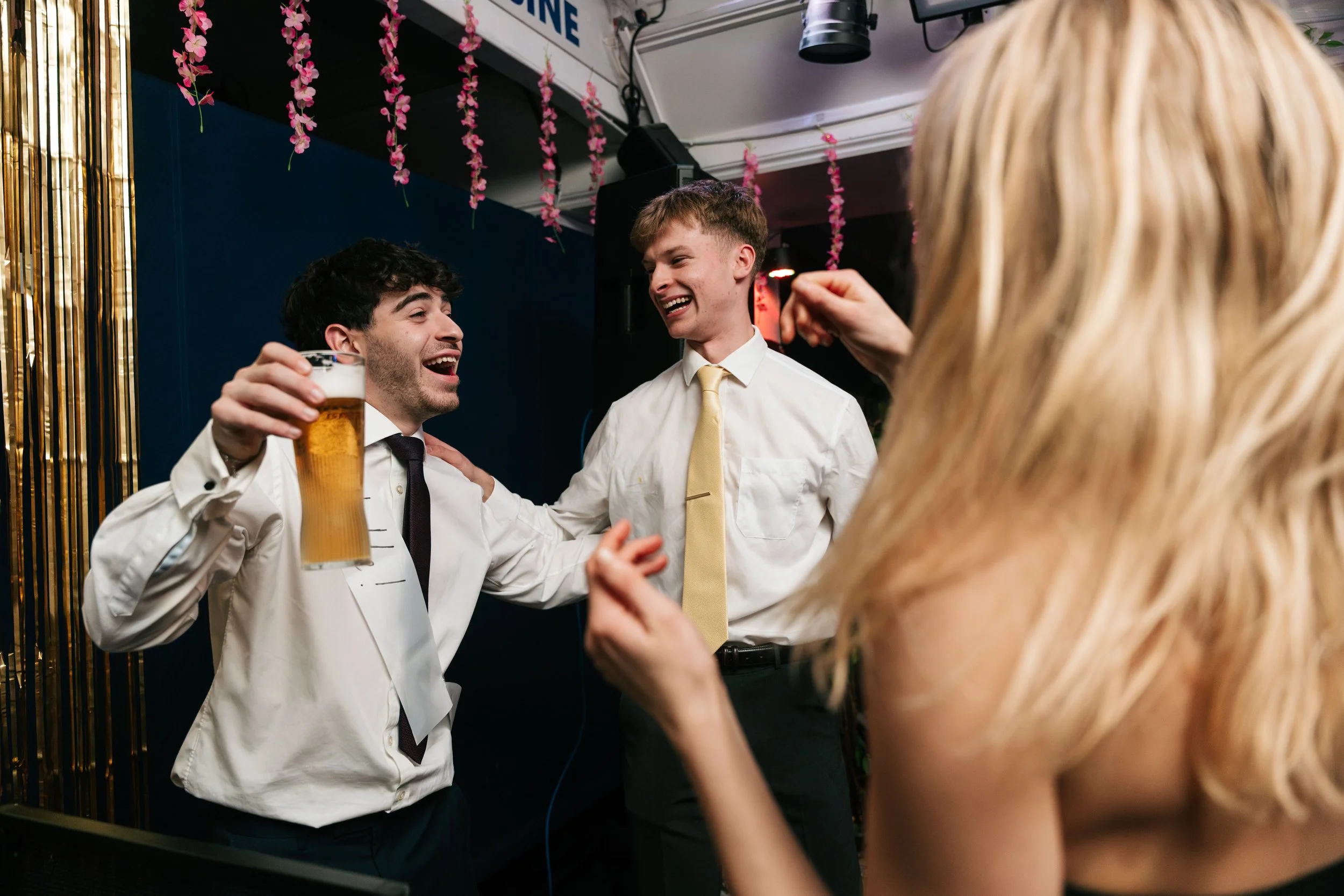 Group of friends dancing and laughing at a party, one holding a drink, in a decorated indoor venue.