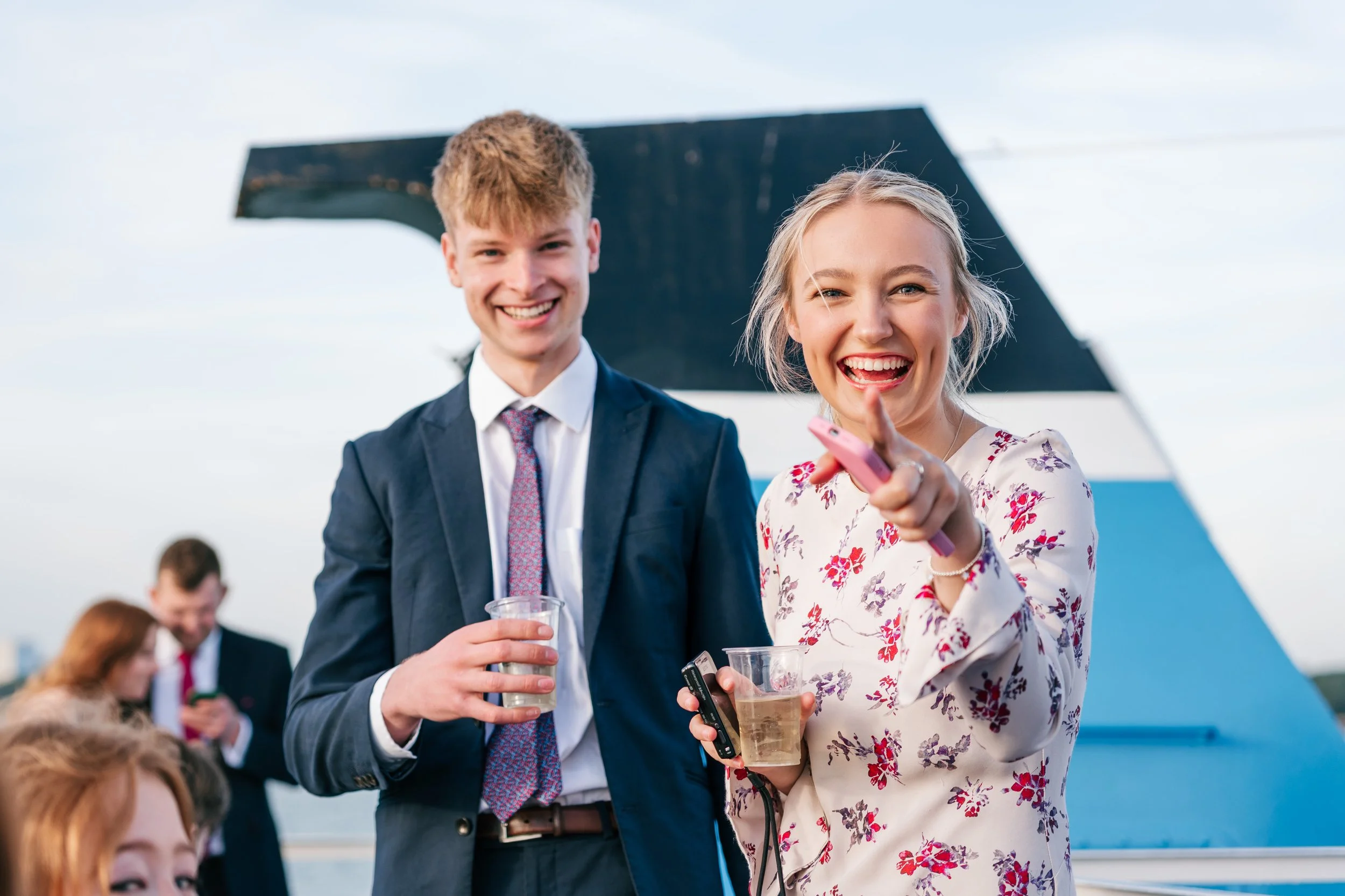 Two young adults at an outdoor event, smiling and holding drinks, with a modern abstract building in the background.