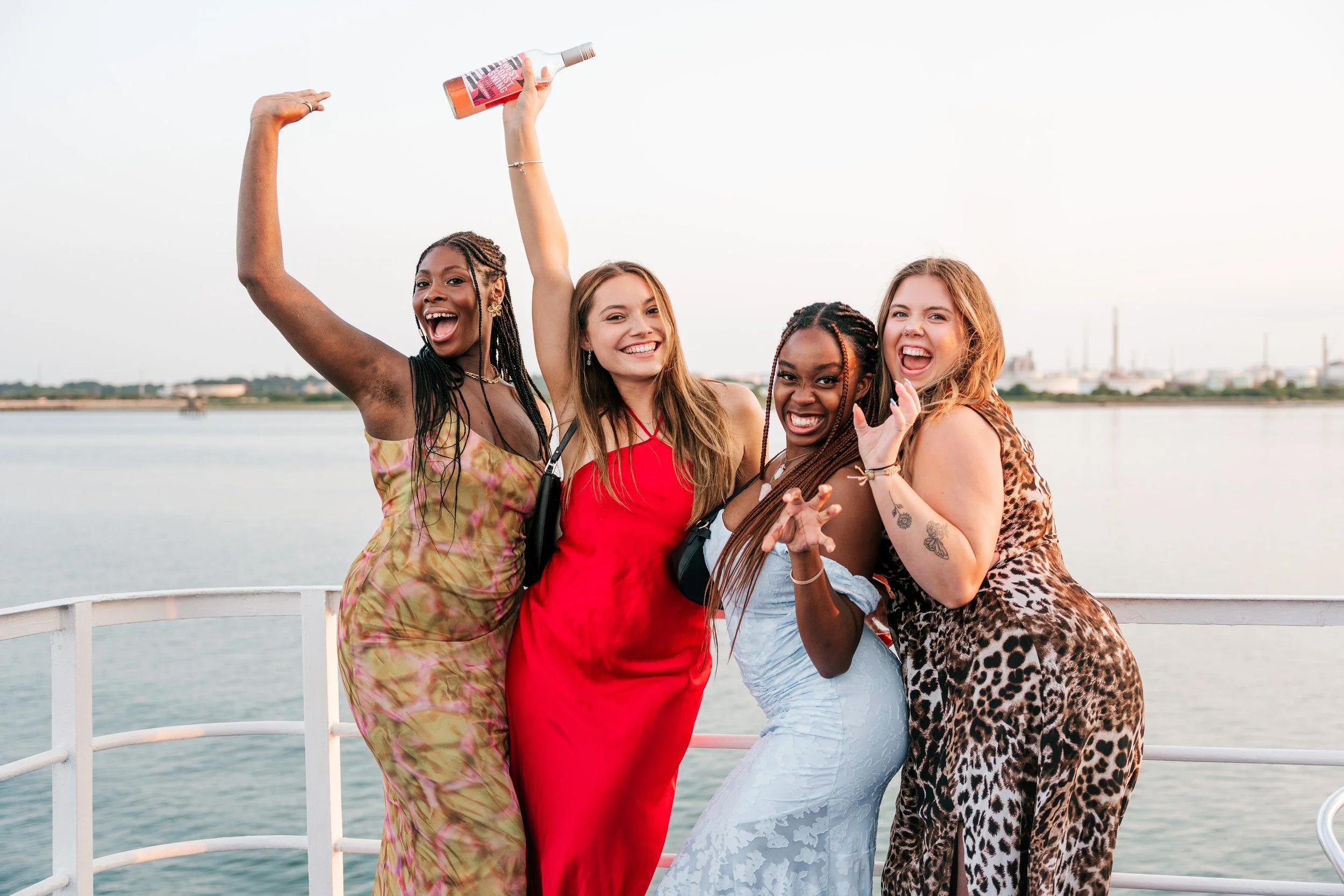 Four women celebrating on a boat with water and industrial buildings in the background, smiling and posing for a photo.