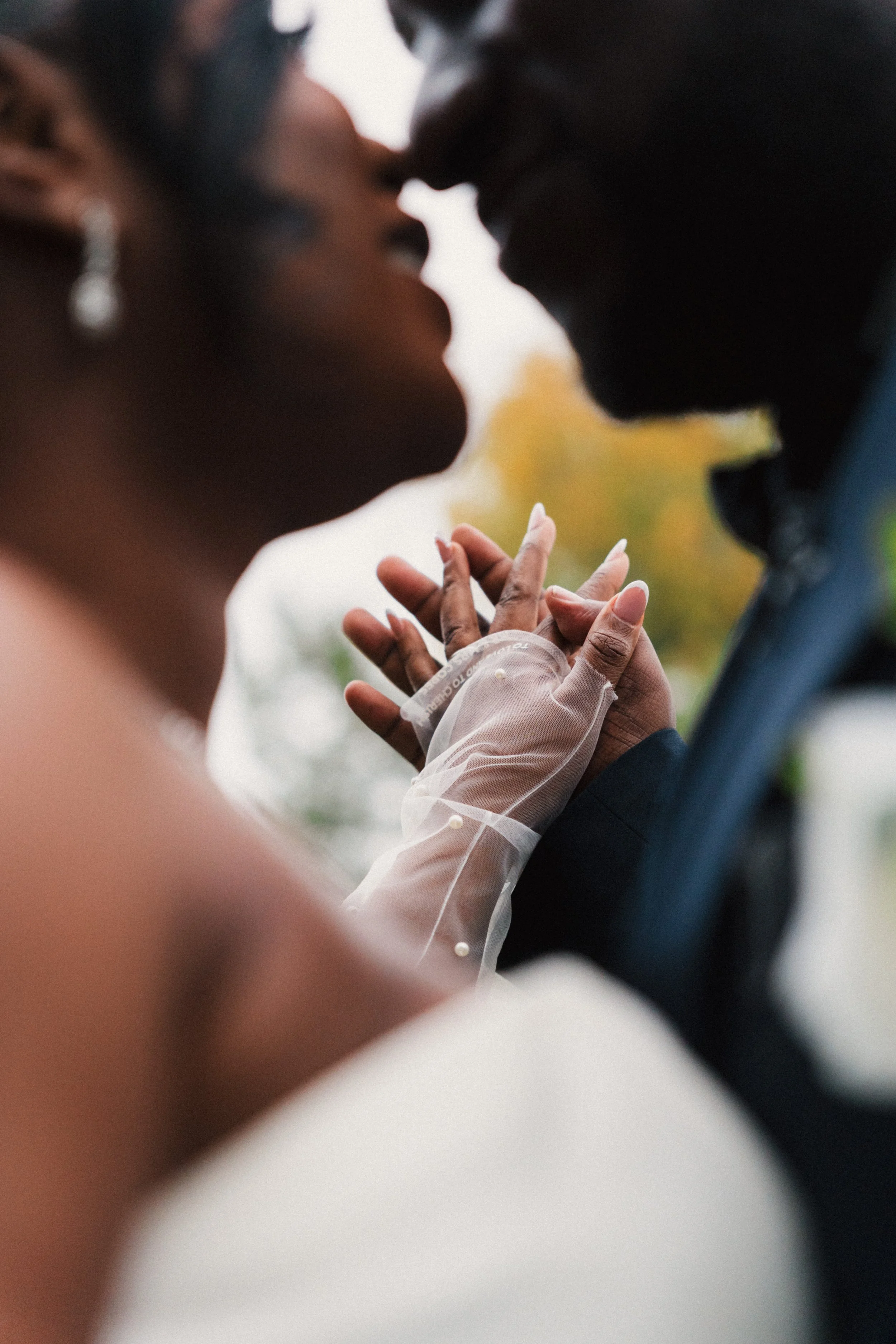 A couple holding hands while sharing a kiss, with the woman's hand featuring a wedding or engagement ring and a sheer glove with pearl embellishments.