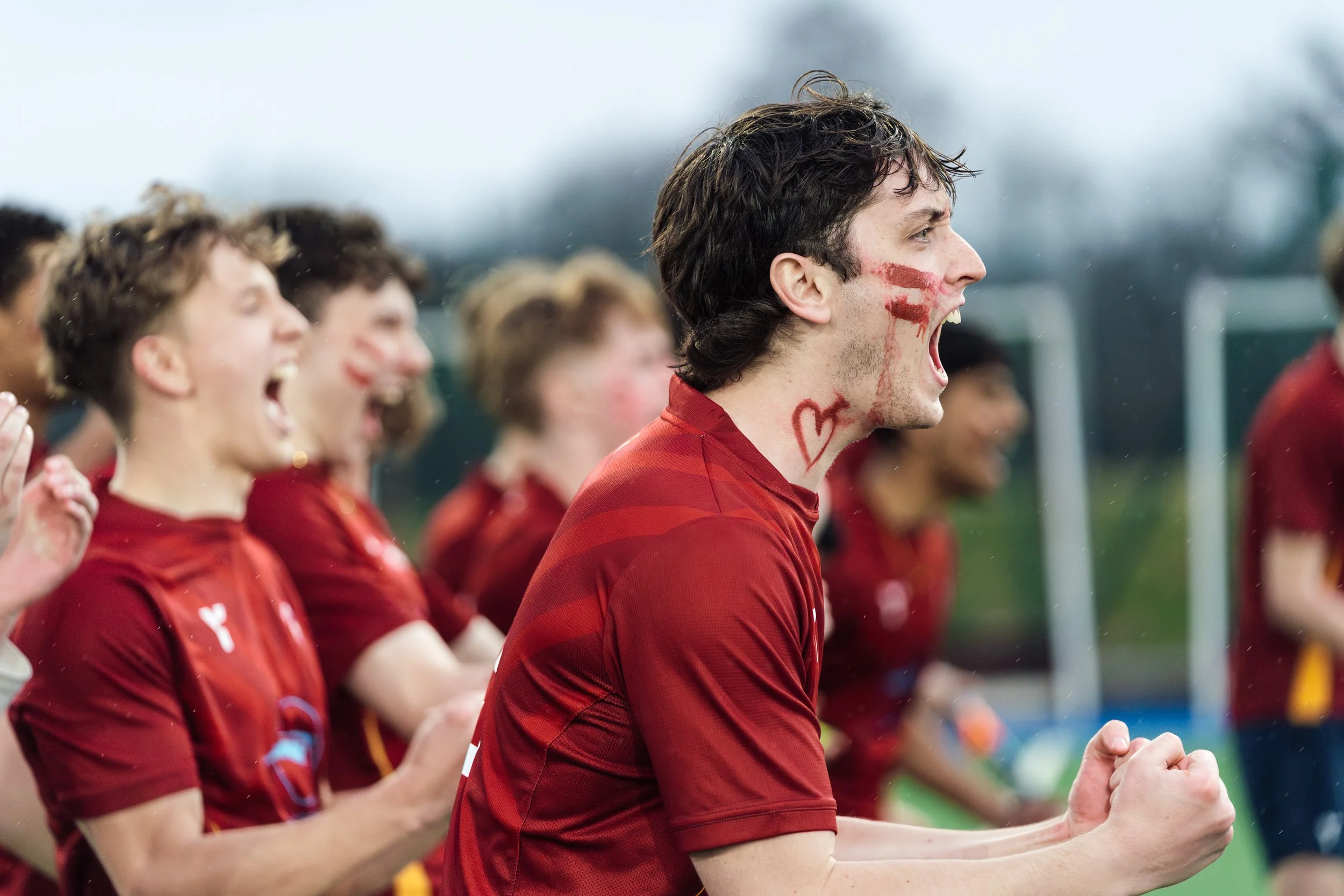Soccer players in red uniforms celebrating on the field, with one player in the foreground with face paint and a heart drawn on his neck, cheering and clenching fists.