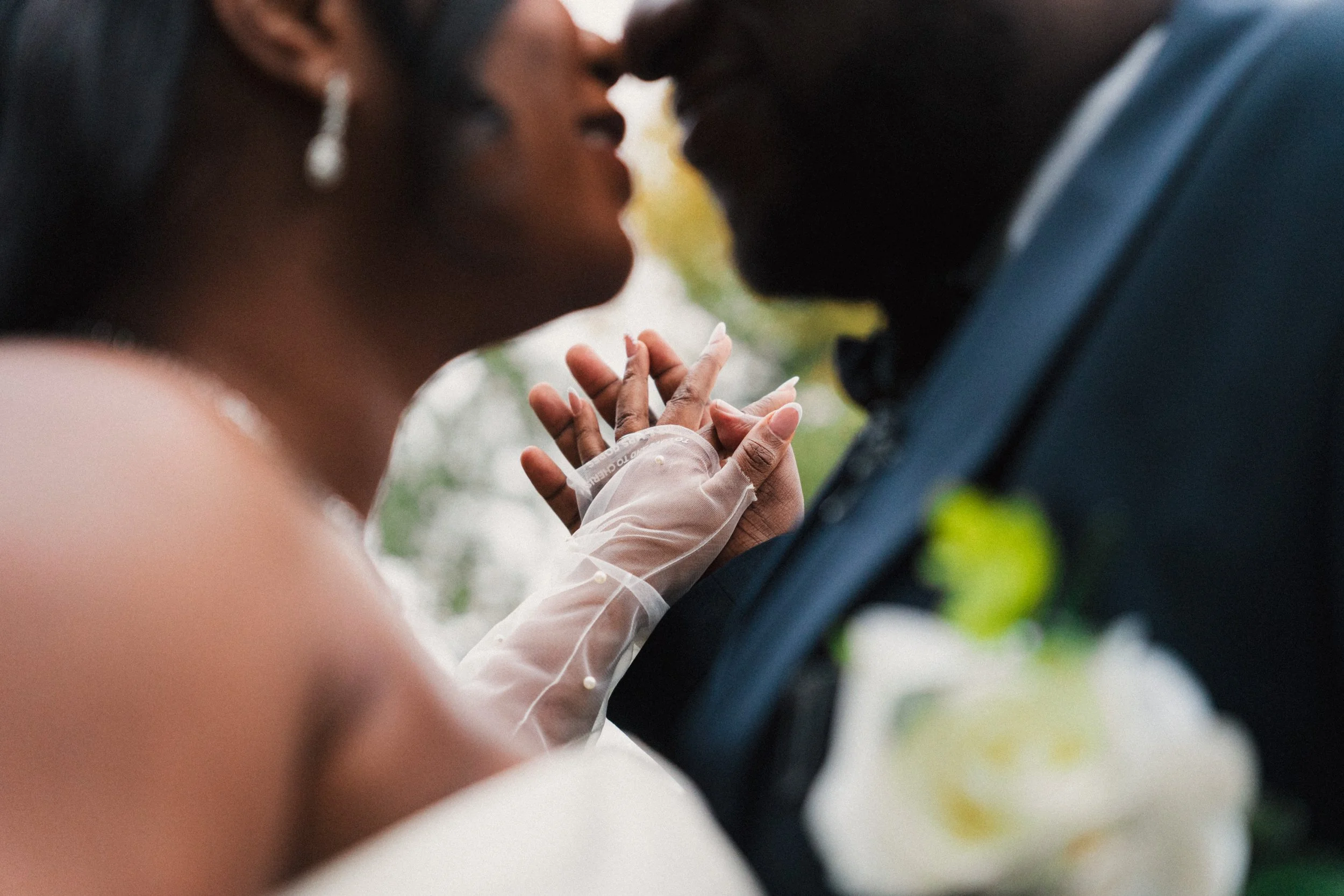 Close-up of a bride and groom touching foreheads during their wedding, holding hands with wedding rings, with a blurred background of trees and white flowers.