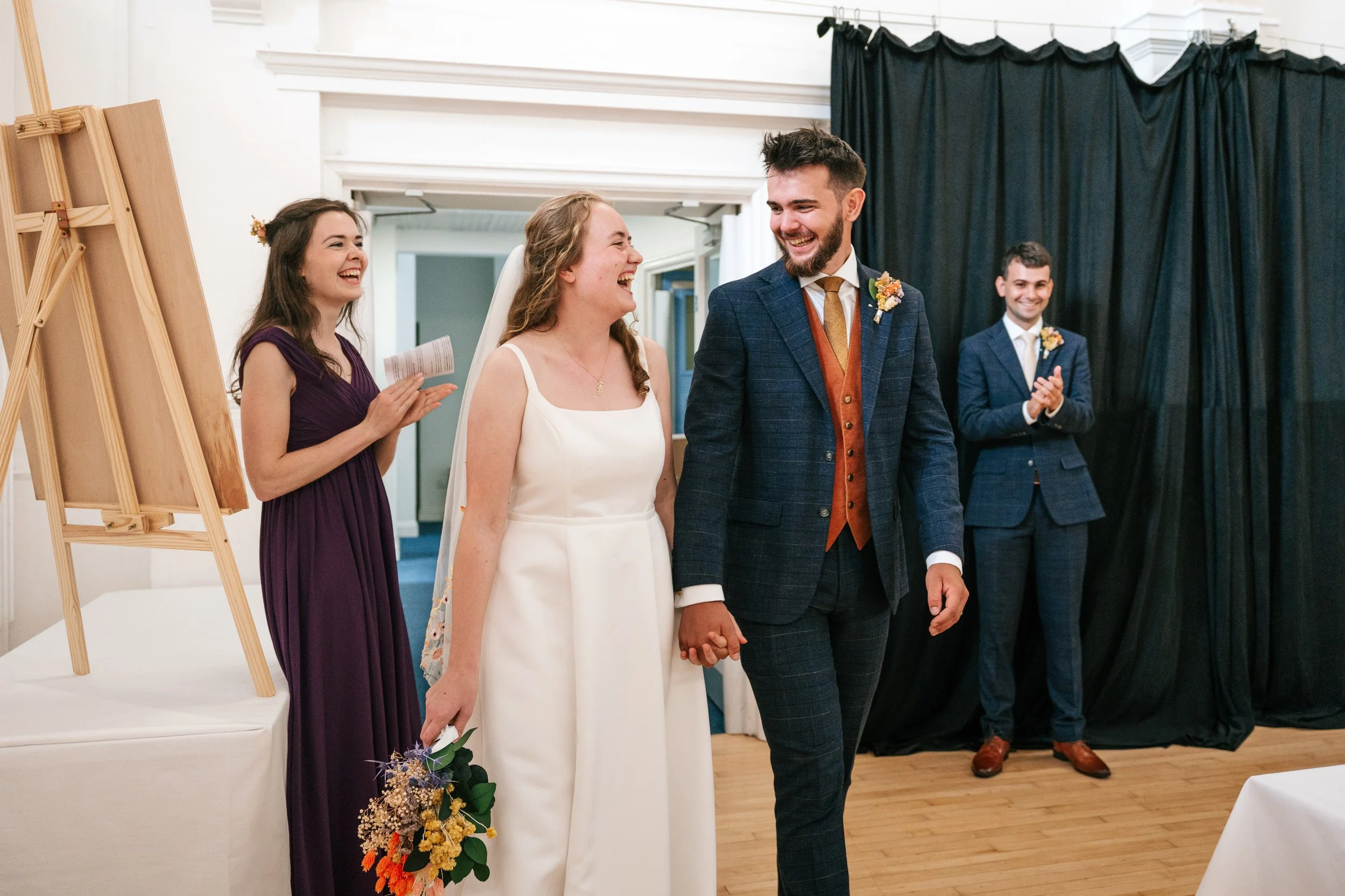 Bride and groom holding hands and smiling during their wedding ceremony, with two bridesmaids and a groomsman clapping in the background, in a decorated indoor venue.