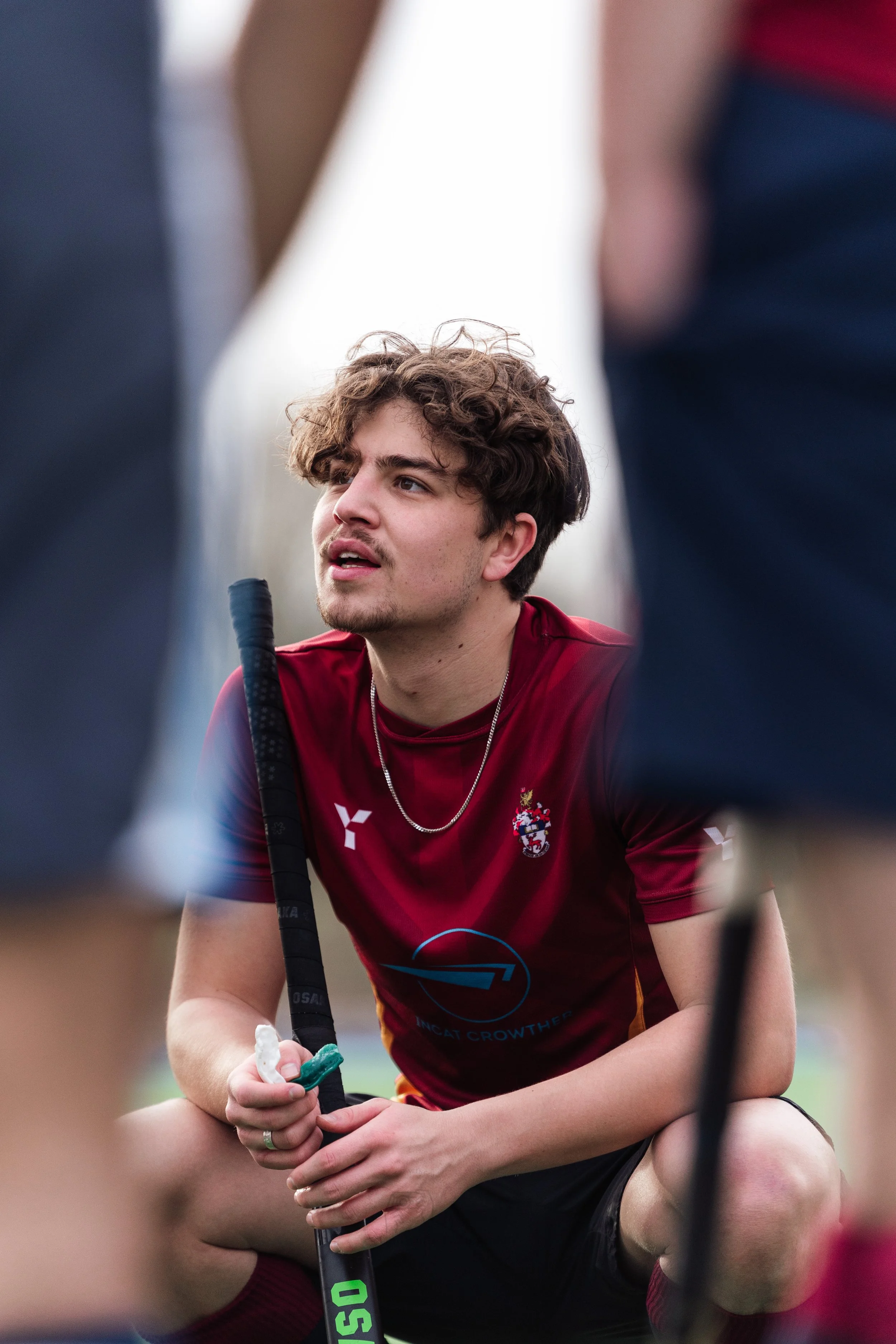 A male field hockey player with curly brown hair, wearing a maroon sports jersey, sitting on the ground and holding his hockey stick, surrounded by other players.