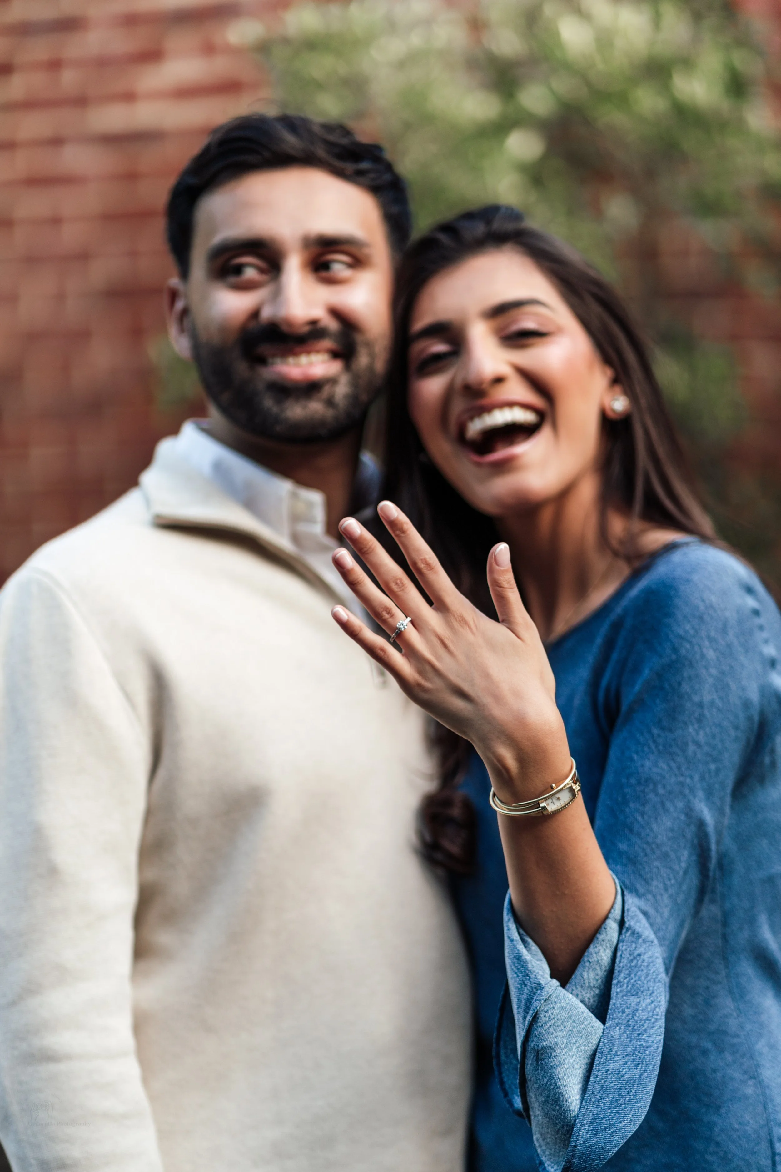 A woman showing her engagement ring with a man smiling next to her in an outdoor setting.